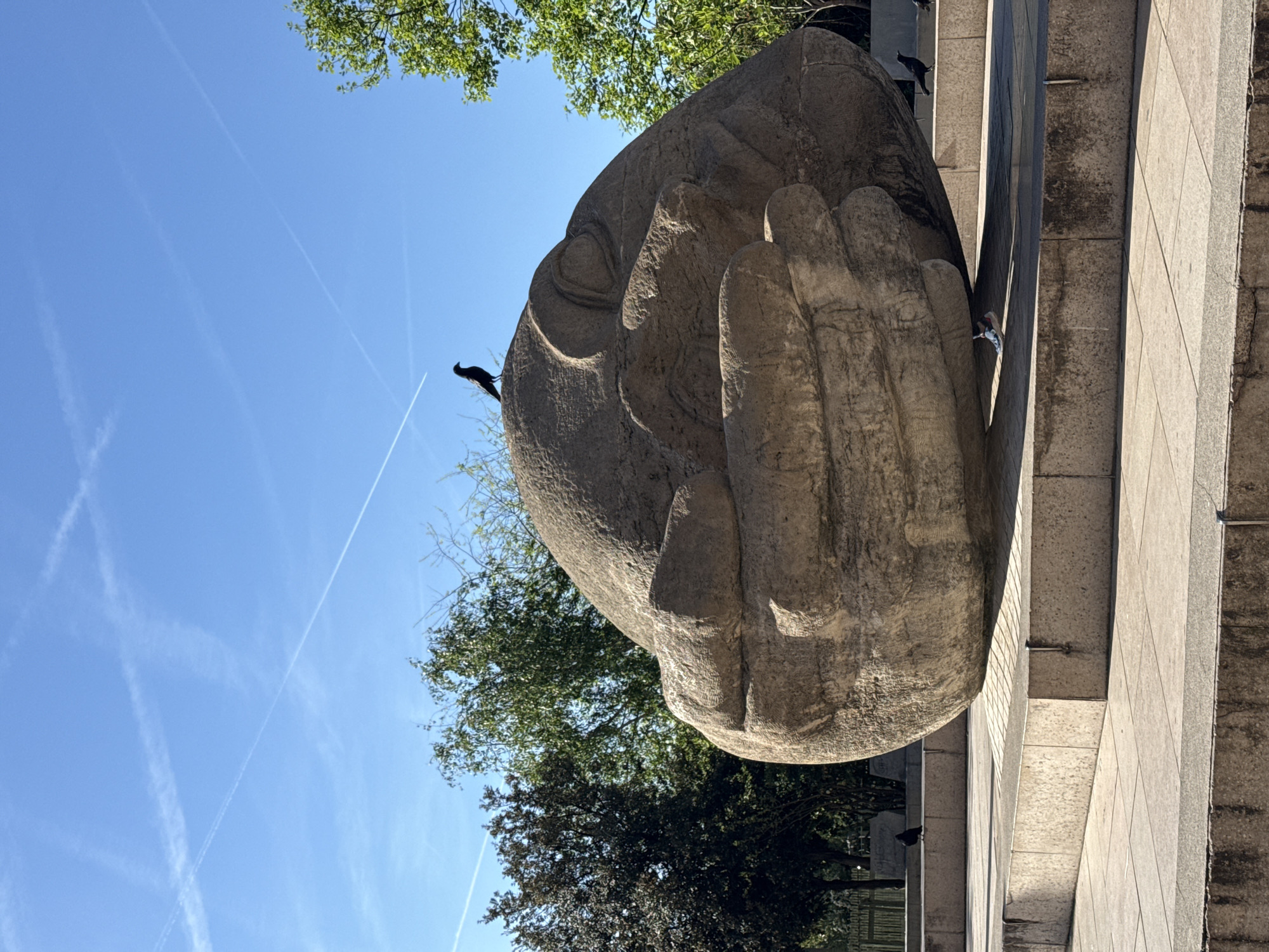 The image shows a large stone sculpture of a sleeping head and hand, located in a public space with trees and a blue sky visible in the background. A bird is perched on the head of the sculpture. The sculpture appears to be situated on a stone platform or ledge. The presence of contrails in the sky suggests recent airplane activity. Based on the sculpture's style and the general environment, it is likely a public artwork located in Paris, France. The sculpture is made of rough-hewn stone, suggesting it may have been carved in place.