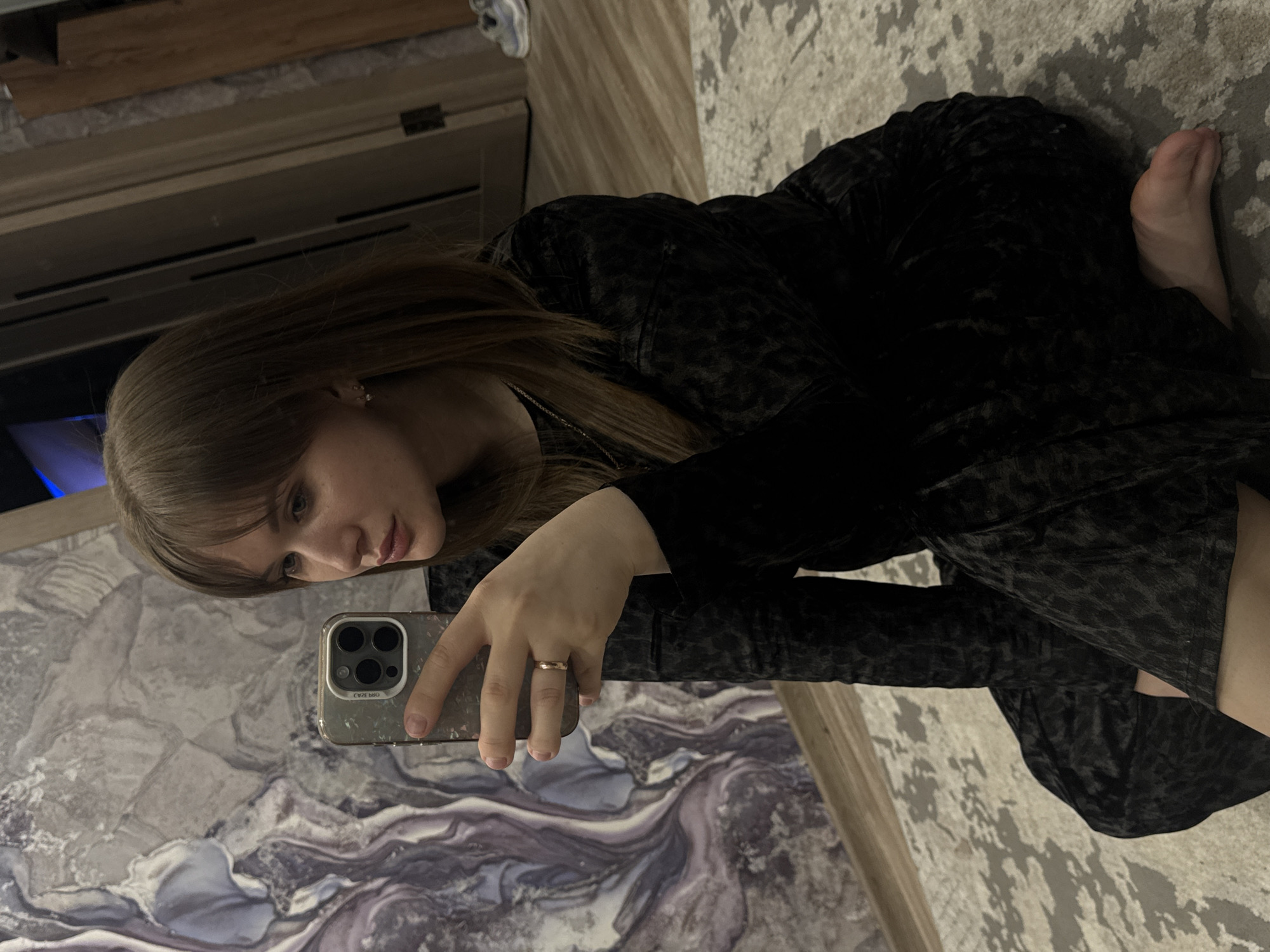 A young woman is depicted taking a selfie indoors, looking down at the screen of her smartphone. She has long, straight, light brown hair, fair skin, and light-colored eyes, with a neutral and focused expression. She is wearing a dark, possibly black or very dark grey, long-sleeved top or dress made of a textured fabric, perhaps crushed velvet or velour, with a subtle patterned design. A thin chain necklace and small stud earrings are visible, and she wears a simple gold-toned ring on her left ring finger. Her bare right foot is also visible.
The woman appears to be kneeling or crouching on an indoor surface. The floor beneath her transitions from a grey and white abstract patterned rug or carpet on the right to a more reflective surface on the left, which features swirling patterns of grey, white, and purple, possibly a polished floor or a large decorative mirror on the floor. In the background, dark wooden or paneling structures are visible, along with a faint blue light source, suggesting ambient indoor lighting. The smartphone she holds in her left hand has a multi-lens camera array and is encased in a clear or lightly textured case. Faint, inverted text or a logo is visible on the phone case, but is indistinct. The scene takes place in Chaikovsky, Russia.
