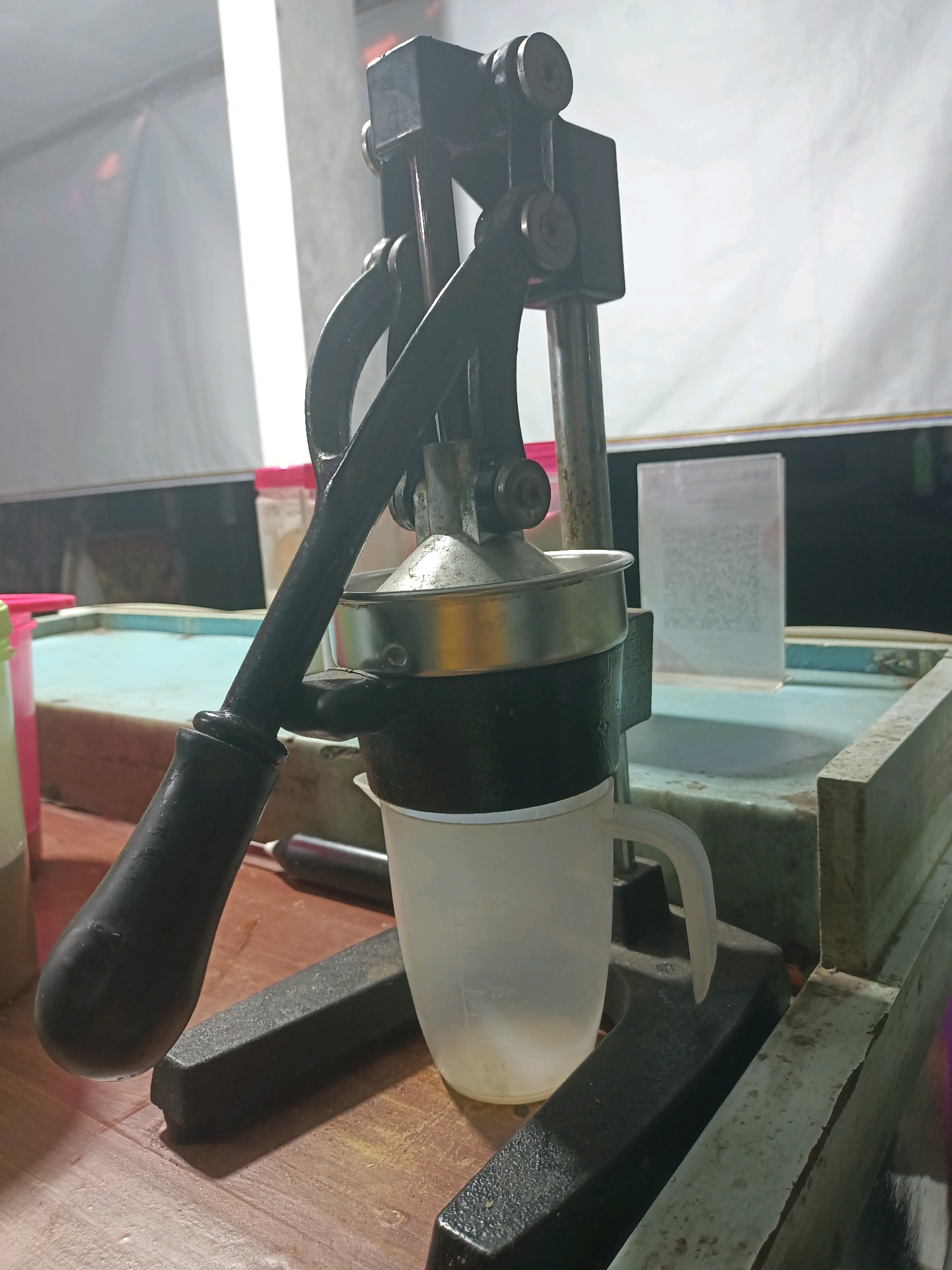 The image shows a manual citrus juicer positioned on a wooden table. The juicer is made of black metal with a silver metal straining bowl. A clear plastic measuring cup with volume markings is placed beneath the juicer to collect the juice. In the background, there is a light-colored tarp, a blue surface that appears to be a table or counter, and a QR code displayed on a stand. The overall setting suggests a food stall or a similar commercial establishment, possibly in Badas, Indonesia, given the context. The lighting is somewhat dim, with a bright light source illuminating the background from the left. There are no people visible in the frame. The scene is static, with no apparent action taking place.