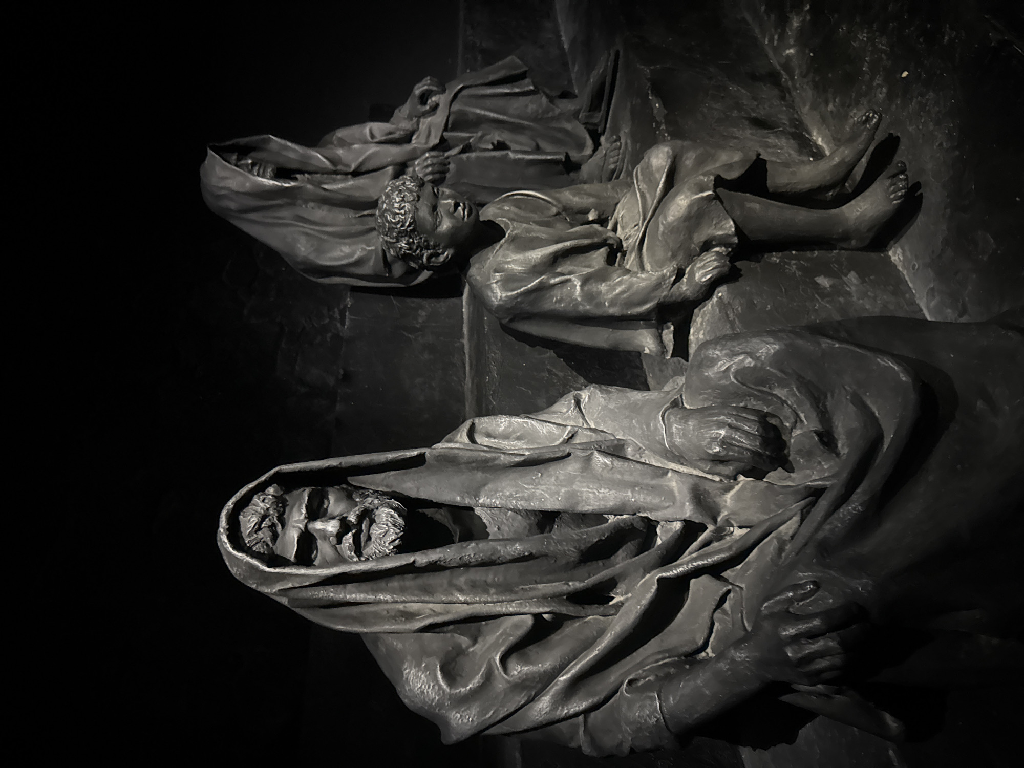 A dimly lit, monochromatic photograph shows a sculpture of three figures, likely a family, seated on stone steps. The figures are draped in heavy, dark robes that obscure most of their bodies. The central figure, a man, is seated on the left, his face visible beneath a hood, with a beard and weathered features. To his right, a child sits, their face turned slightly away. Behind them and to the right, a third figure, cloaked and shrouded, is also seated, their features obscured. The setting appears to be an indoor or shadowed outdoor space, with the lighting focused on the sculptures, creating deep shadows and highlighting the textured surfaces of the stone. The sculpture's style is realistic, conveying a sense of somberness or contemplation. It is located at the Lod Airport in the Tamar Regional Council, Israel.