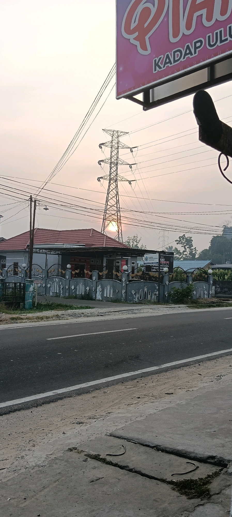 The image depicts a roadside scene in Kuala Kapuas, Indonesia, during what appears to be sunrise or sunset, given the low angle of the sun and the soft lighting. A prominent high-voltage transmission tower stands in the middle ground, with its silhouette against the hazy sky. The sun is partially obscured by the tower structure, creating a lens flare effect.
In front of the tower, a single-story building with a red-tiled roof is visible, bordered by a gray, decorative fence. The building has several windows and a covered outdoor area. A sign with text, possibly advertising a business, is mounted on the fence.
The foreground of the image is dominated by a paved road with white lane markings. The road surface is asphalt. To the right of the road, a concrete sidewalk or pathway is present, with a section that appears to be a manhole cover or drainage grate. Two dark, slender objects, consistent with the appearance of snakes, are lying on the concrete surface near the grate.
The overall environment suggests a semi-urban or suburban setting, with infrastructure like power lines and a main road adjacent to residential or commercial properties. The sky is overcast or hazy, contributing to the diffused light.