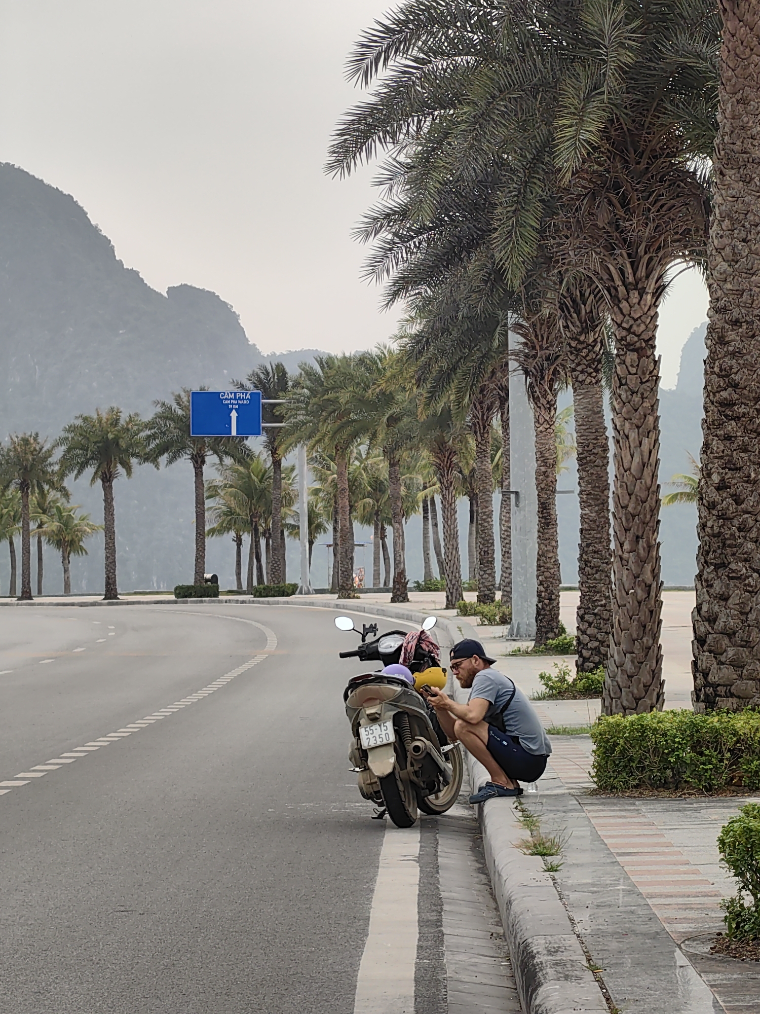 A man is crouched beside a parked motorcycle on the side of a road in Ha Long Ward, Vietnam. The motorcycle is tan in color with the license plate "55-Y5 2350." The man is wearing a grey t-shirt, dark shorts, a black baseball cap, glasses, and blue shoes. He is facing the motorcycle and appears to be focused on something in his hands. The road is paved with a white dashed line down the center. Palm trees line the road, and mountains are visible in the background. A blue road sign is visible in the distance.