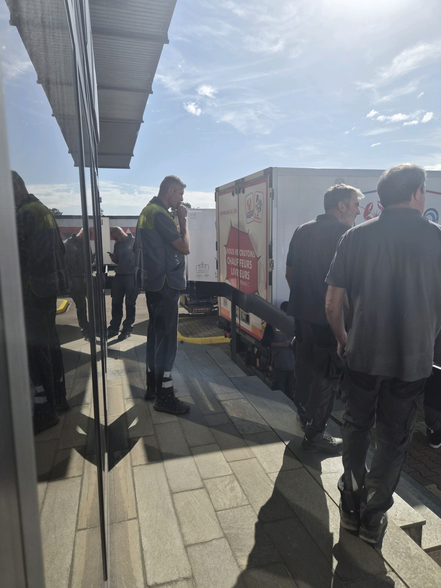 In Leudelange, Luxembourg, under a bright, sunny sky with scattered clouds, a group of men in work attire are gathered around a white delivery truck. The scene appears to be a commercial or industrial area, with a modern building featuring reflective windows on the left side of the frame, mirroring the sky and the people. The ground is paved with light-colored rectangular tiles.
Several men are visible, most of whom are wearing dark work pants with reflective strips and dark shirts or high-visibility vests. One man in the foreground, facing right, is wearing a blue and yellow high-visibility vest over a dark shirt and dark pants. He is standing with his hand near his mouth, possibly smoking or deep in thought.
In the background, other men are seen near the truck. One man is looking at his phone, while others are standing by the side of the truck. The truck itself is white with some branding and text. Visible text on the truck includes "NOUS RECRUTONS CHAUFF EURS LIVR EURS EUROS" and "PROVENÇALE.LU/JOB". There are also images of crustaceans on the truck.
The time of day is clearly daytime, indicated by the strong sunlight and distinct shadows. The overall mood seems professional and operational, with the workers engaged in their tasks or taking a brief pause. The setting suggests a logistics or delivery operation in progress.