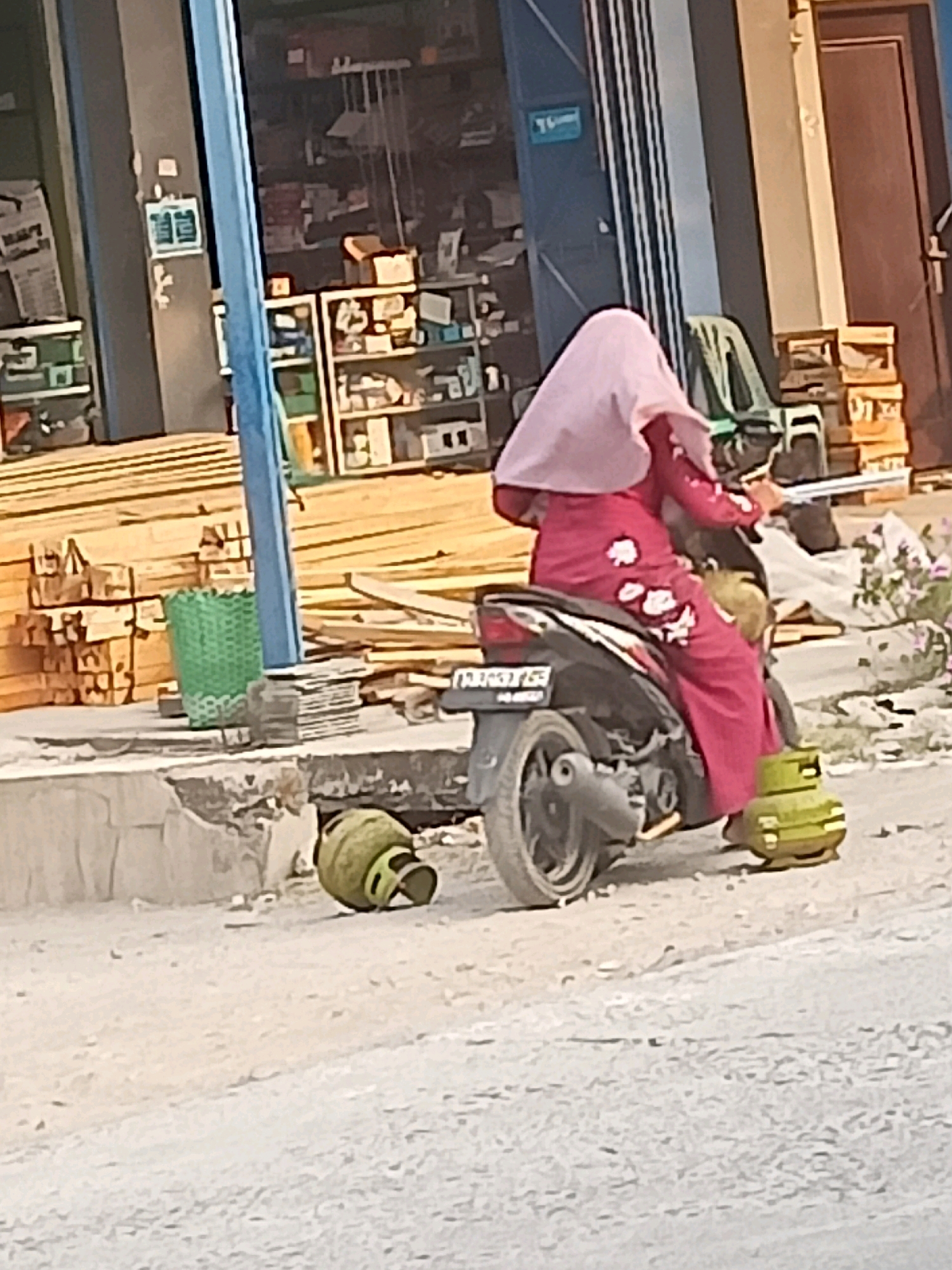 In Karangtalun, Indonesia, a person wearing a pink headscarf and a patterned pink dress is seated on a scooter. The scooter is parked on a dirt road next to a building that appears to be a shop or warehouse. Two green gas cylinders are visible on the ground; one is upright near the rear of the scooter, and the other is lying on its side. In the background, stacks of lumber are visible, along with shelves stocked with various items inside the building.