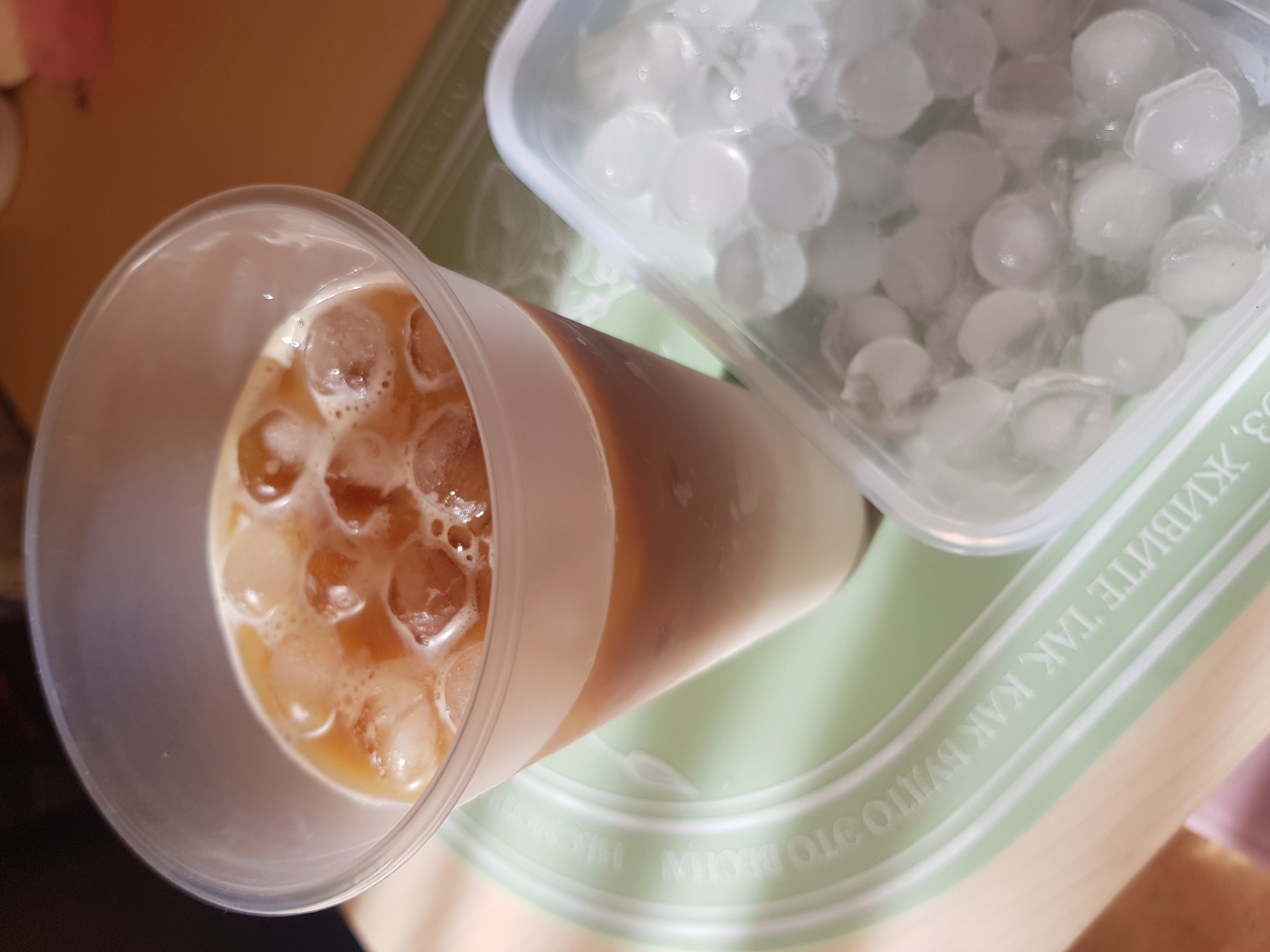 A high-angle shot shows a plastic cup filled with a tan liquid and spherical ice cubes, alongside a clear container overflowing with similar ice spheres. These items rest on a light green surface with what appears to be white text or designs, positioned on a beige or light wood tabletop. The setting suggests an indoor location. The city and specific details for a location are not discernible from the image.