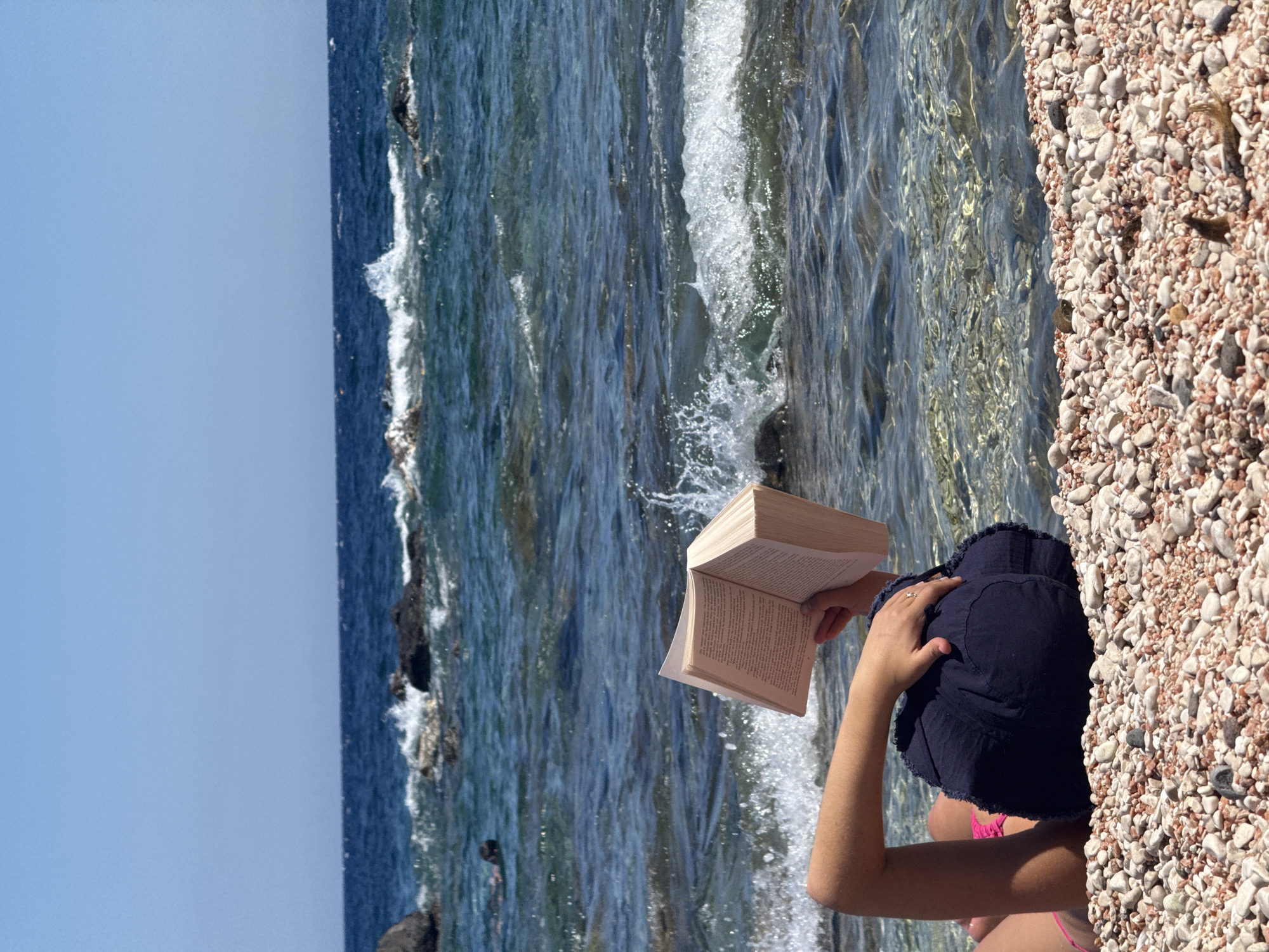 In Cala Gonone, Italy, a person lies on a pebble beach, reading a book. The person is wearing pink bikini bottoms and dark blue shorts, with a blue bucket hat on their head. Their right arm is raised, holding the open book. The sea is calm with gentle waves breaking on the shore, and the sky is clear and blue. The image captures a serene moment of leisure by the Mediterranean Sea.