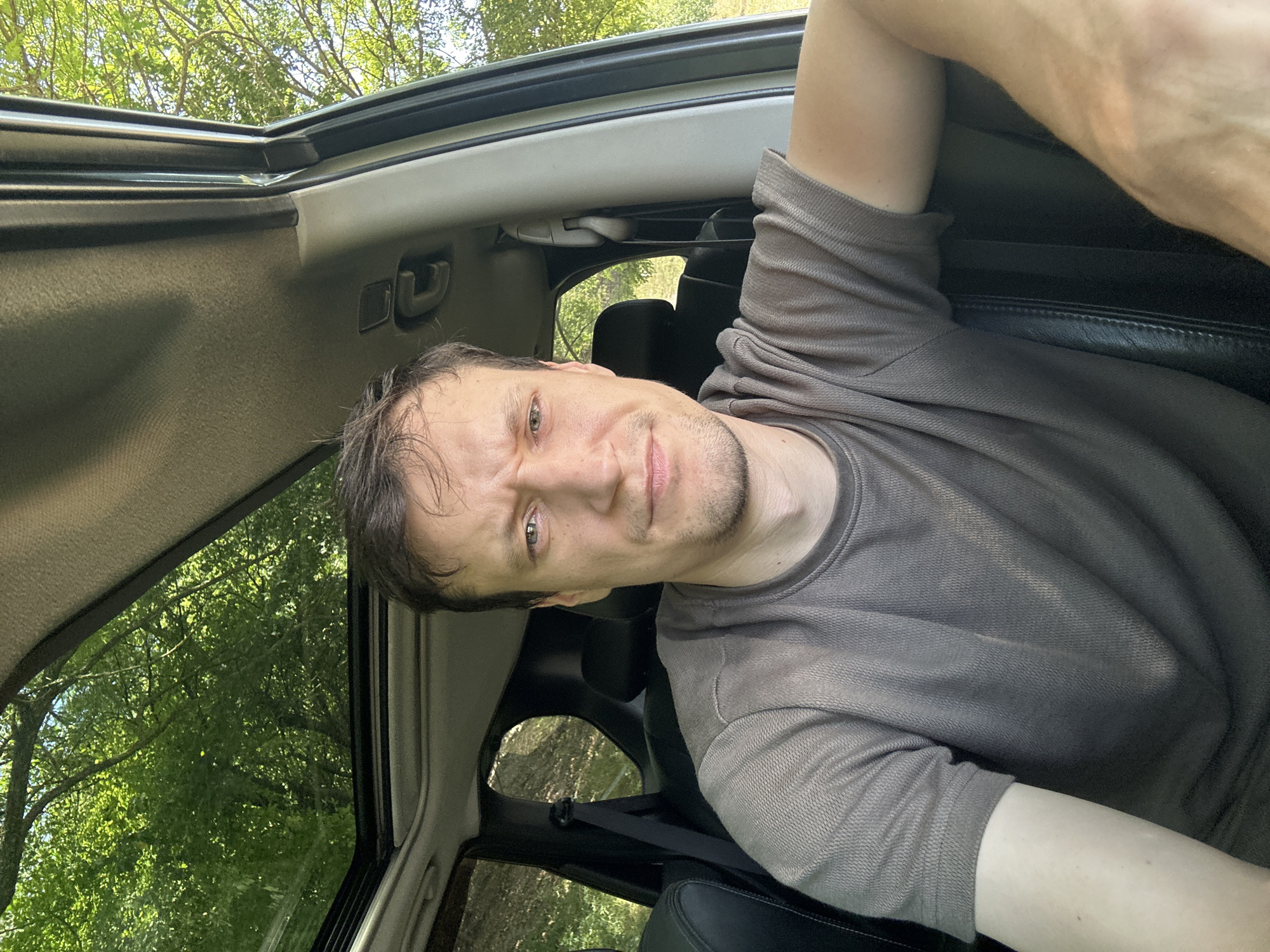 A man is sitting in the driver's seat of a car, looking at the camera. The car has a sunroof that is open, revealing trees and sky. The man is wearing a grey t-shirt. The interior of the car appears to be black leather. The scene suggests the car is parked outdoors, likely in a natural setting such as a forest or park. There are no indications of the specific location in the city of Makiv, Ukraine.