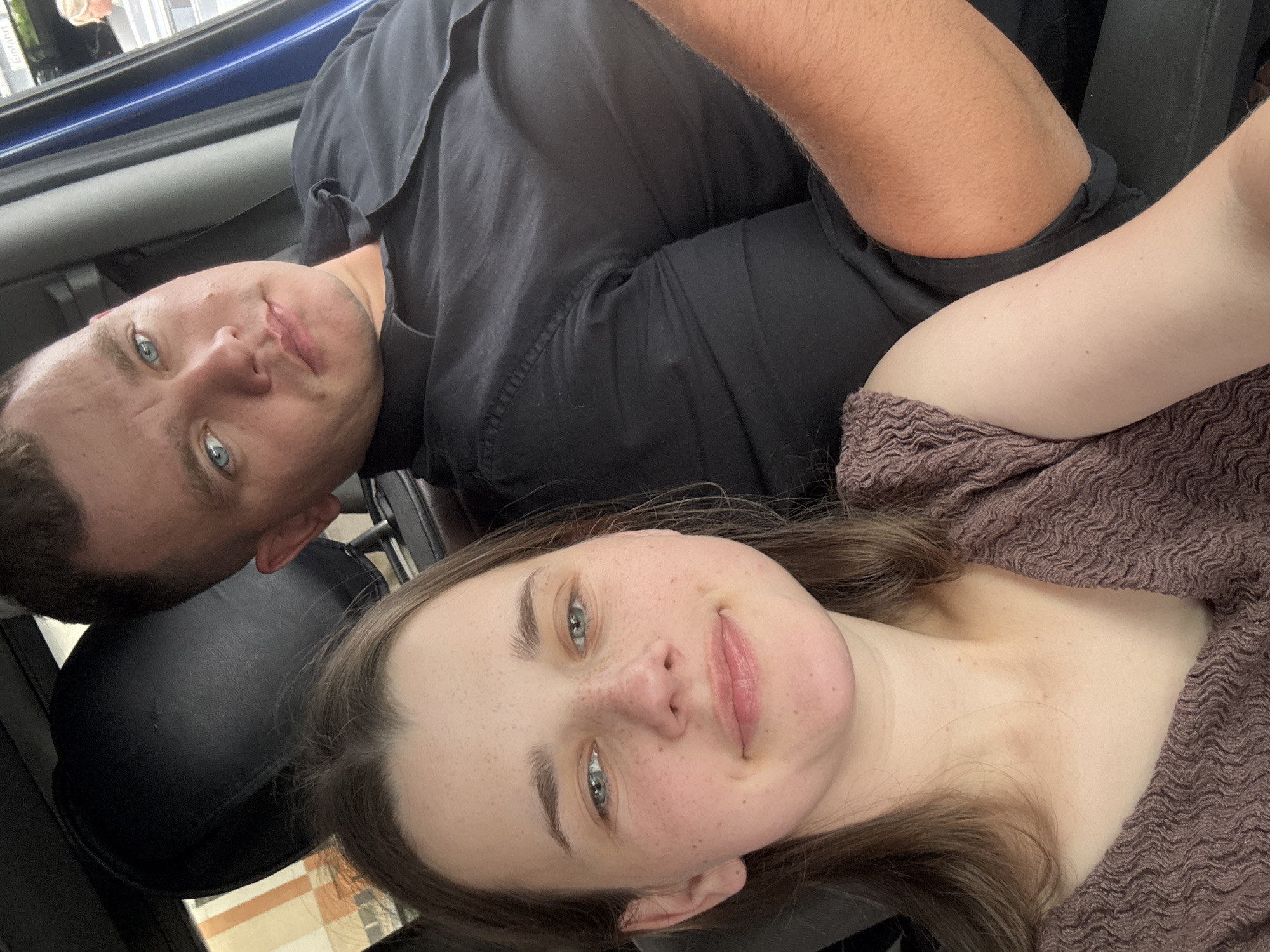 Two people, a woman and a man, are in a car. The woman is in the foreground, facing the camera and smiling. She has light brown hair and is wearing a brown, textured top. She has freckles across her nose and cheeks. The man is in the background, to the right of the woman, also looking towards the camera. He has short brown hair and blue eyes. He is wearing a black collared shirt. The interior of the car is visible, including a black headrest and a blue door panel. Through the window, a building with beige and orange elements is visible, suggesting an urban environment. The overall scene appears to be a casual moment captured within the city of Koblenz, Germany.