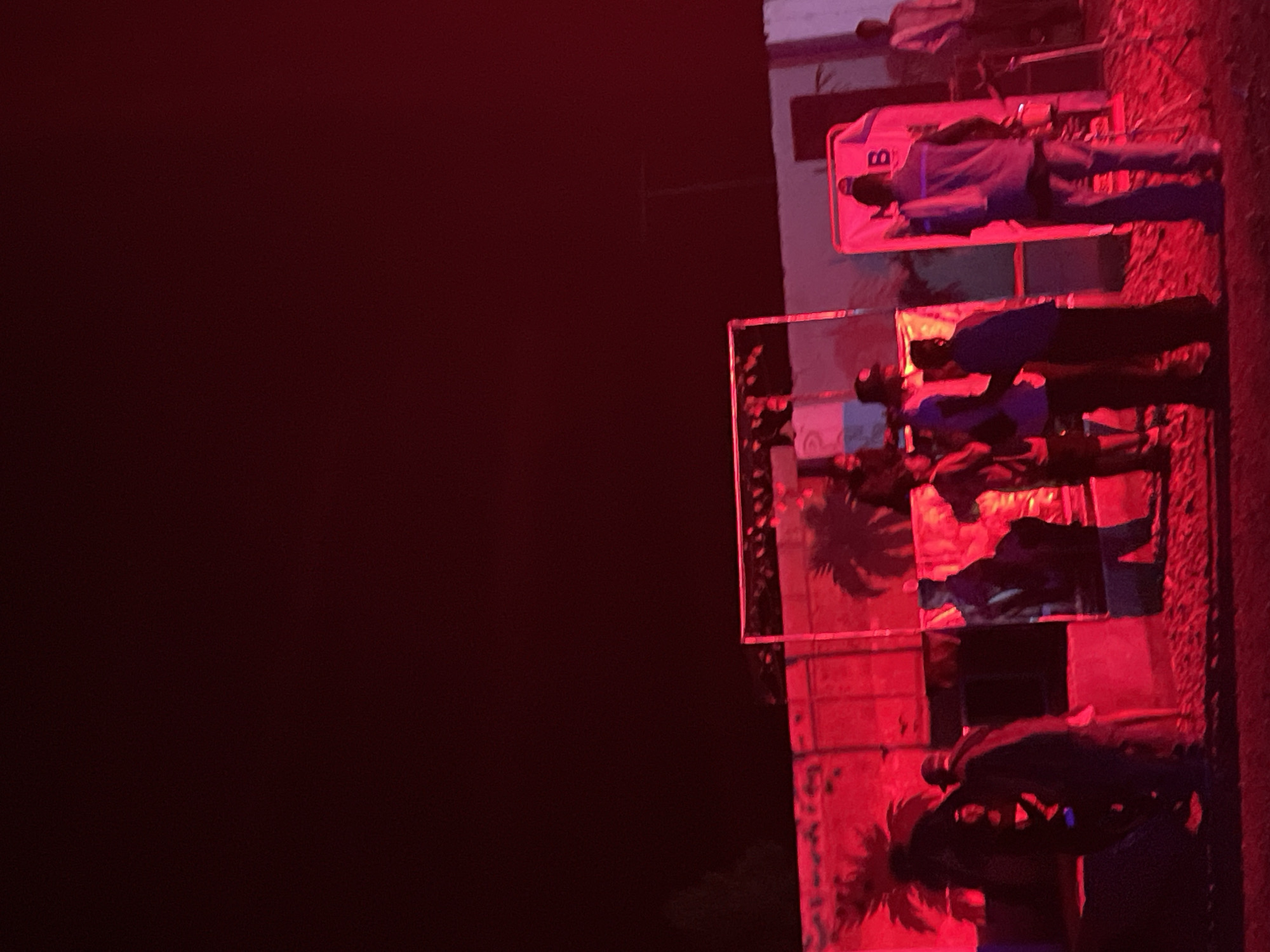 In the city of Durumi, Nigeria, at night, a group of individuals are gathered outdoors under red lighting. The scene appears to be some sort of event or performance setup. In the background, there's a structure with what looks like shipping containers, illuminated by the red lights, with palm tree silhouettes visible against them. In the foreground, a large, reflective structure, possibly a mirror or screen, is positioned. Several people are standing around, some interacting with each other, while others are observing. One individual stands facing away from the camera, wearing a vest over a shirt, and their reflection is visible. Another large object, also lit in pink, has the letters "MB" on it. The ground is uneven, and there are some cables and stands present, suggesting a setup for sound or lighting equipment. The overall atmosphere is dark with vibrant red and pink hues from the lighting.