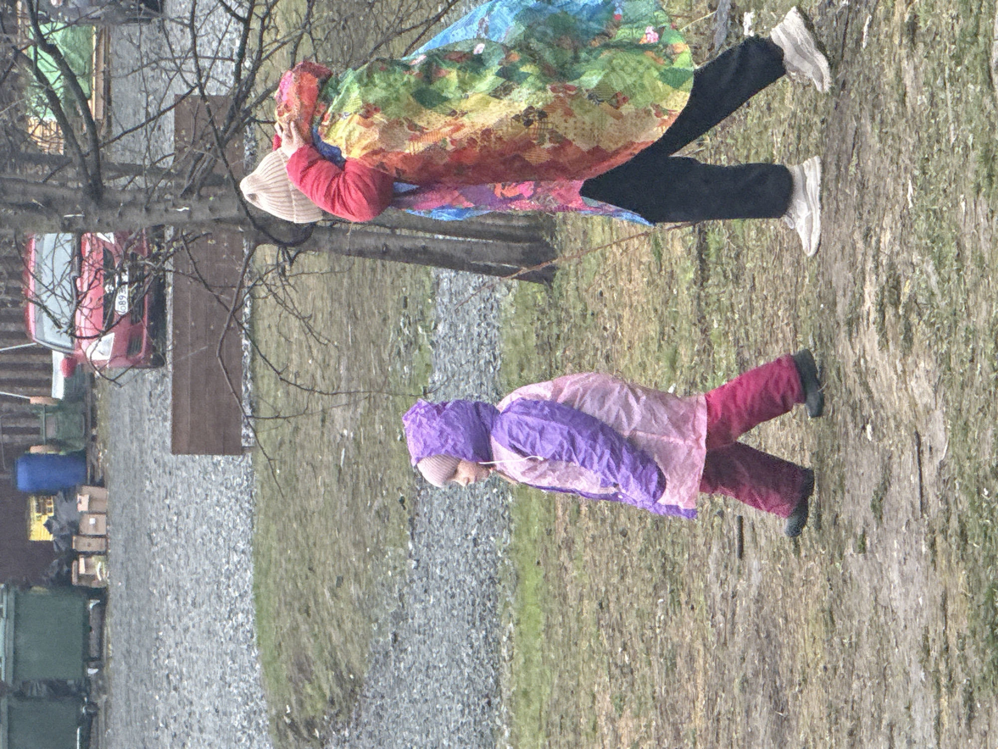 This outdoor image, taken in Yekaterinburg, Russia, depicts two individuals during what appears to be an overcast day in a colder season, possibly late winter or early spring.
In the middle ground, a taller person wears a distinctive, colorful coat, possibly with a patchwork or artistic design, in various shades including red, blue, green, and yellow. They are dressed in a red long-sleeved top, black trousers, and light-colored sneakers, topped with a light-colored beanie. Their face is largely obscured.
Ahead and to the right, a shorter individual, likely a child, is prominent. This person is dressed in a two-toned purple and pink hooded jacket, dark pink or maroon trousers, and dark boots. They also wear a light-colored beanie and appear to be in mid-stride, looking forward or downward.
The setting features a mix of terrain: a wide area of grey gravel occupies the left side of the ground, contrasting with a patch of sparse, dry, and muddy grass on the right. Bare tree branches are visible in the upper left, reinforcing the sense of a colder time of year. In the background, to the left, a red car is parked, with some numbers like "089" partially visible on its license plate. Nearby, various objects including a blue barrel, cardboard boxes, and other items are scattered, hinting at a utilitarian or semi-rural environment. The diffused daylight suggests a cloudy sky. No clear activity beyond walking or standing is discernible, and no specific emotions are evident.