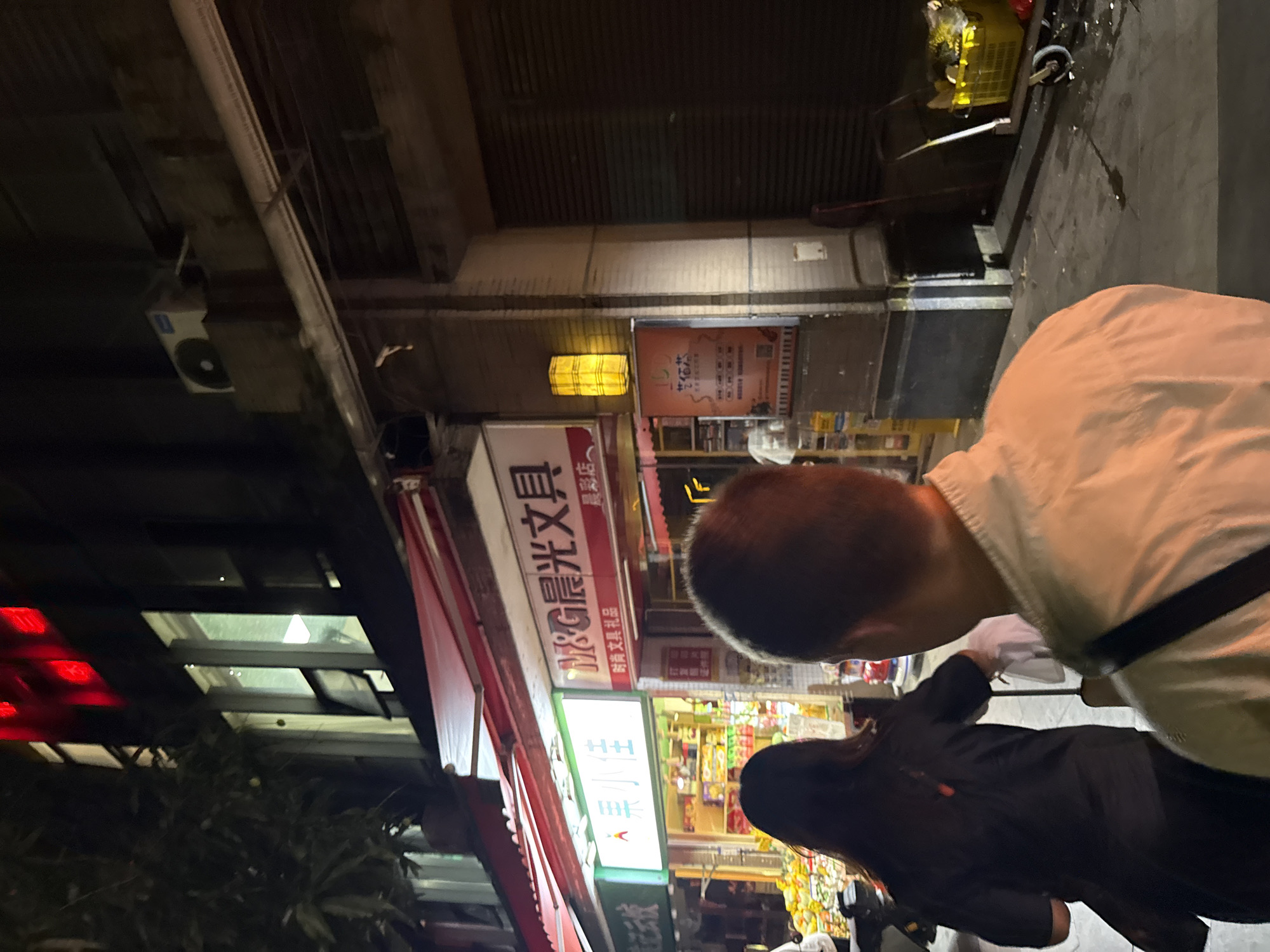 The image shows a nighttime street scene in what appears to be an urban area. The focus is on two individuals, one wearing a light-colored shirt and a dark strap, and the other a person with long hair wearing a black jacket or coat. They are walking along a sidewalk past a storefront, which is adorned with red awnings and illuminated signage in Chinese characters. The shop seems to be a stationery or office supply store. Other shops and buildings are visible in the background, and a street light provides illumination. The image does not provide enough details to ascertain the specific location in Haizhu District, China.