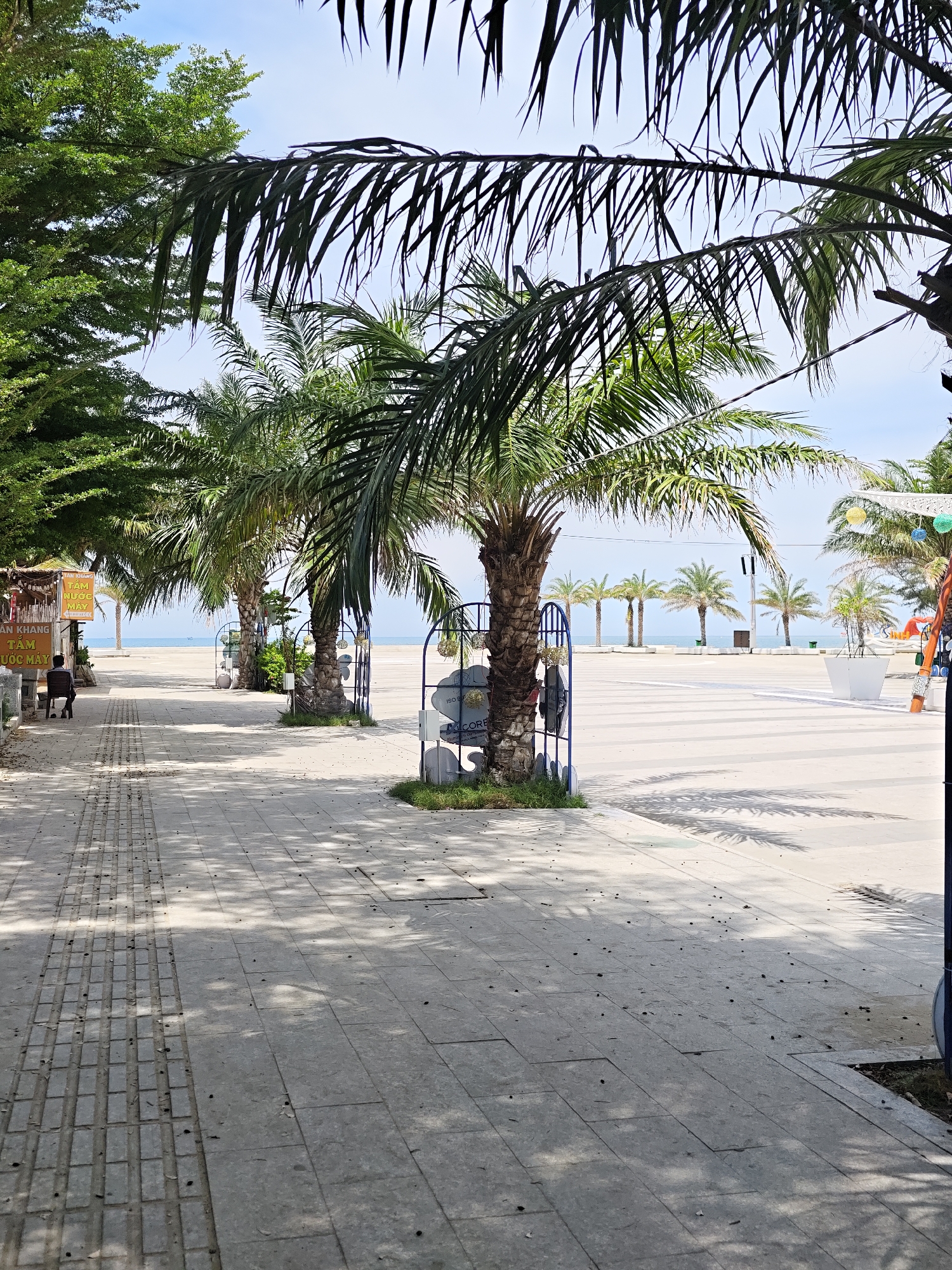 This image shows a sunny day on a wide, paved walkway near a beach in Phan Rang – Tháp Chàm, Vietnam. Several palm trees line the walkway, with their fronds casting dappled shadows on the ground. In the background, the calm sea is visible under a clear blue sky. A few people are present, including one person sitting on a chair under a stall on the left side of the walkway. There are also decorative fences and planters along the path. The overall scene suggests a tranquil and scenic seaside location.