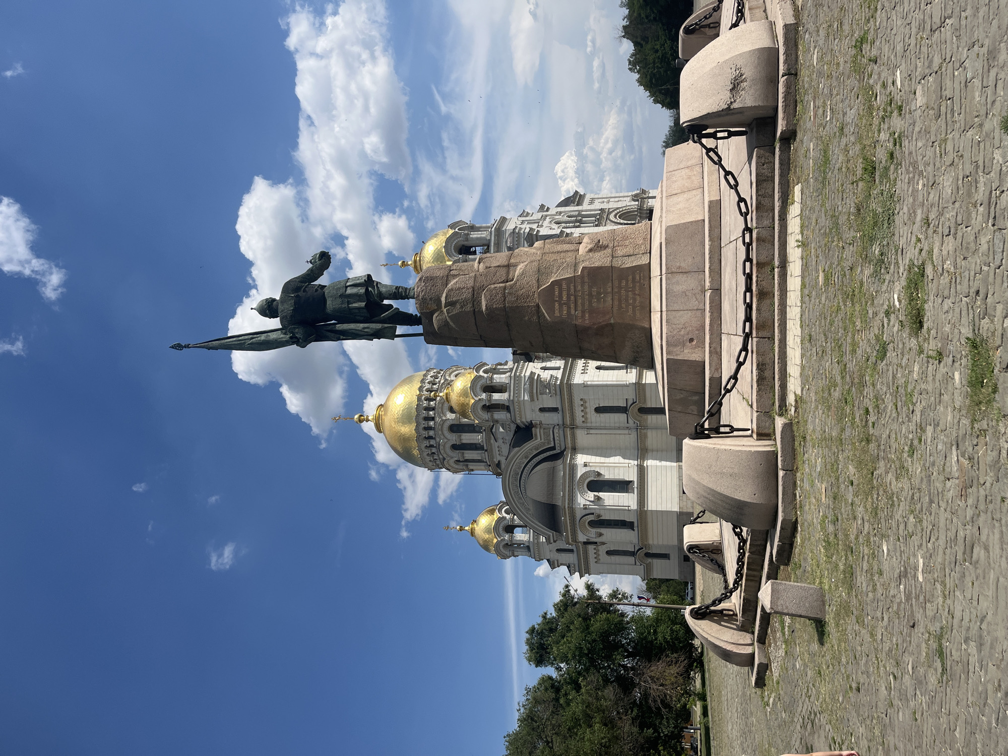 The image depicts the Ascension Cathedral in Novocherkassk, Russia, a prominent landmark with a striking white exterior and ornate golden domes. In front of the cathedral stands a large bronze statue of a man holding a flag, mounted on a stone pedestal. The statue faces away from the viewer, looking towards the cathedral. The area surrounding the monument and cathedral is paved with cobblestones and features a large stone base with heavy chains, suggesting it's a historical or memorial site. The sky is a clear blue with scattered white clouds. Lush green trees are visible to the left of the cathedral, and more trees form a distant treeline on the right. A sliver of a person's arm and leg is visible on the far left edge of the frame. The overall scene conveys a sense of grandeur and historical significance.