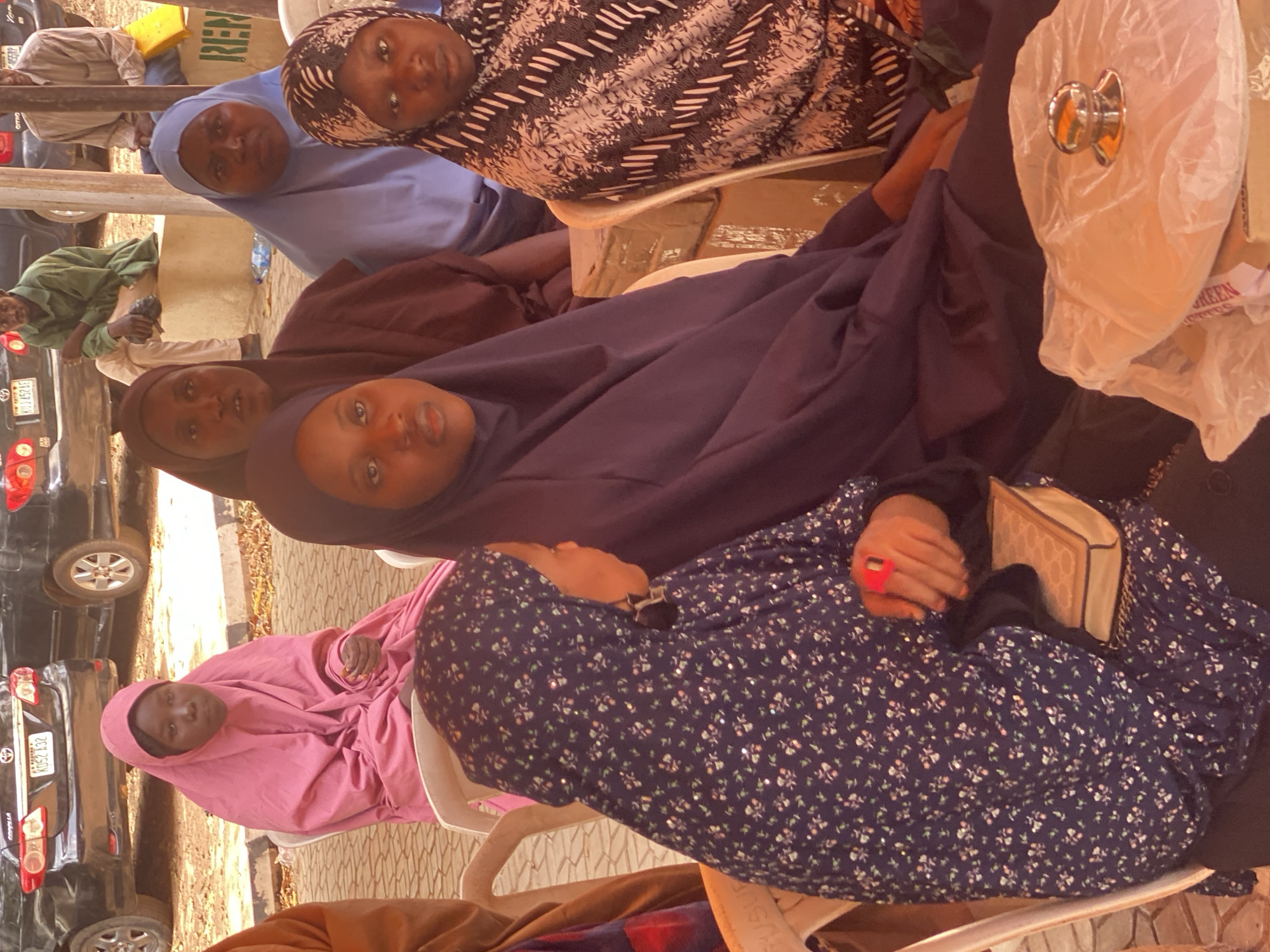 The image shows several people, primarily women, seated outdoors in Unguwar Rimi, Nigeria. They appear to be attending a gathering. All women are wearing hijabs and/or abayas of different colors and patterns: pink, brown, blue, black and white. Some of the women are sitting on plastic chairs. There are cars parked nearby. One of the women is holding a small purse. A white plastic bag with what appears to be a metal dispenser on it is visible on a table.