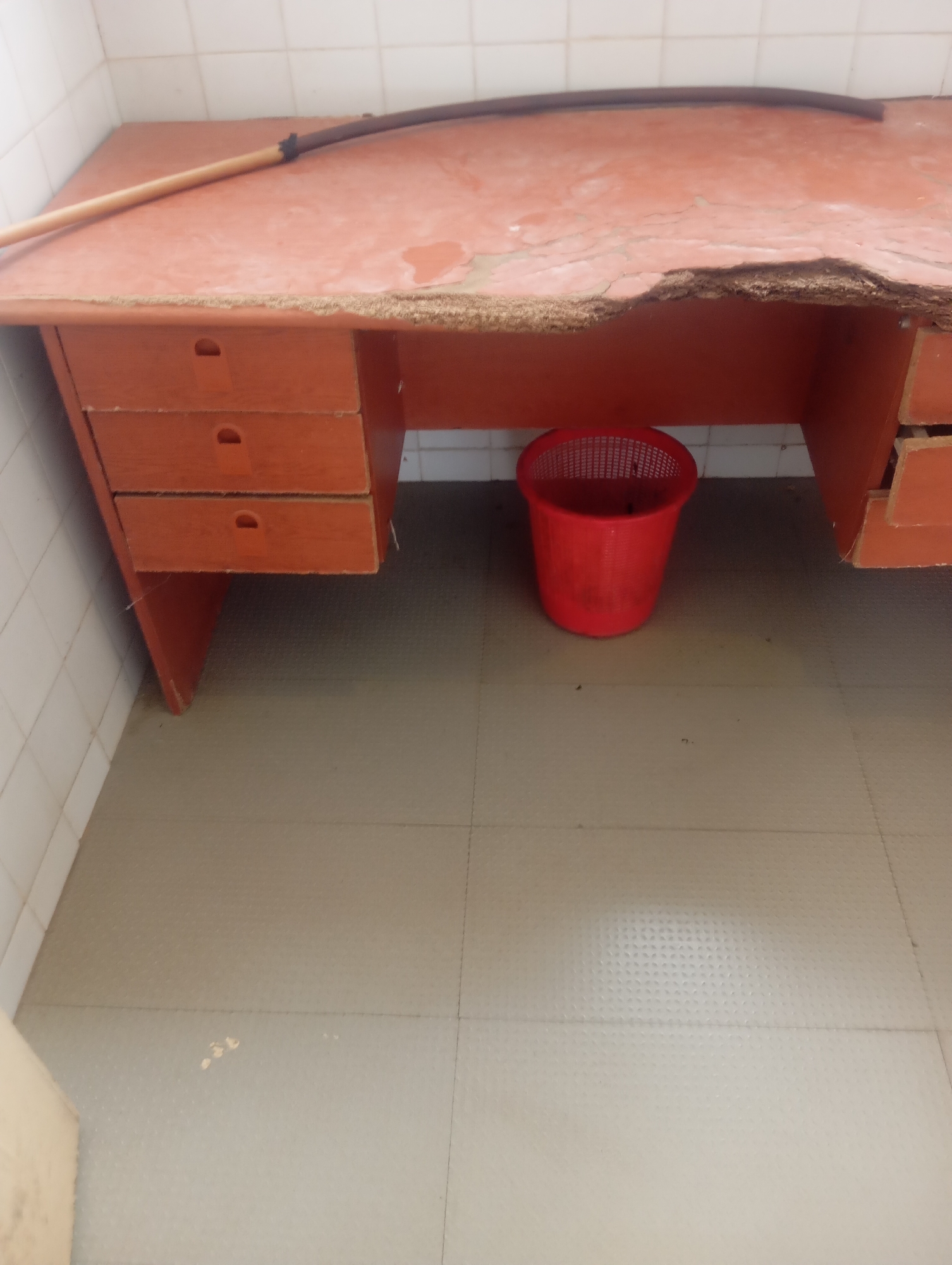 The image shows a simple wooden desk with orange drawers against a tiled wall. A broomstick rests across the desk, which appears to be old and worn, with a damaged edge. Beneath the desk, a small red plastic waste bin is visible on a tiled floor. The scene is consistent with a room in Jos, Nigeria, likely a workshop or a storage area, given the utilitarian nature of the objects and the condition of the desk. There are no people or animals present in the image. The lighting is somewhat dim, suggesting an indoor environment without direct sunlight. The overall impression is one of disuse or neglect.