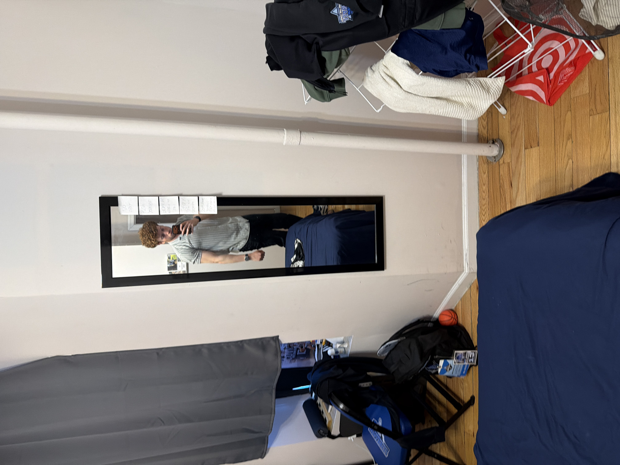 A bedroom scene. A man with curly hair is reflected in a black-framed mirror, holding a phone as if taking a picture. Six small pieces of paper are taped above the mirror. A gray curtain hangs on the left. A blue-covered bed takes up the right of the frame. A clothes drying rack with items is near the top. A basketball sits on the floor near a black backpack. The setting appears to be a room in New York City, judging by the building's interior architectural details, although visual markers confirming the specific city are absent.