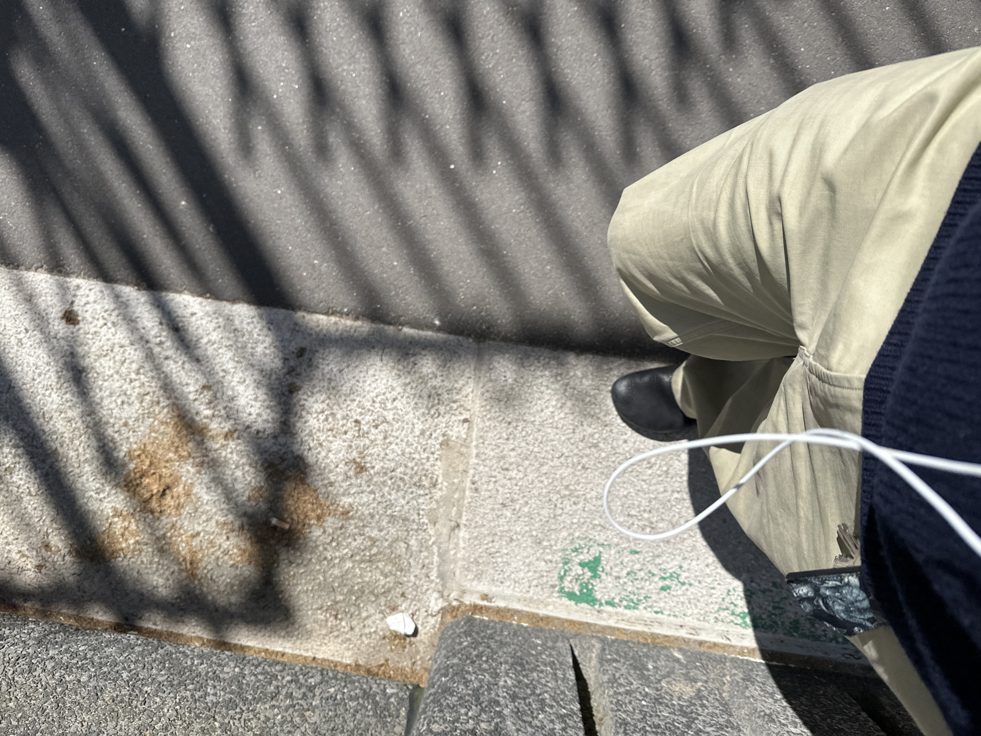 The image shows a person standing on a sidewalk in London, United Kingdom. The sidewalk is made of concrete and stone. The person is wearing beige pants, a navy blue sweater, and black shoes. They are holding a white cable, possibly for earbuds or headphones. Sunlight filters through trees, casting shadows on the sidewalk. A cigarette butt sits on the curb.