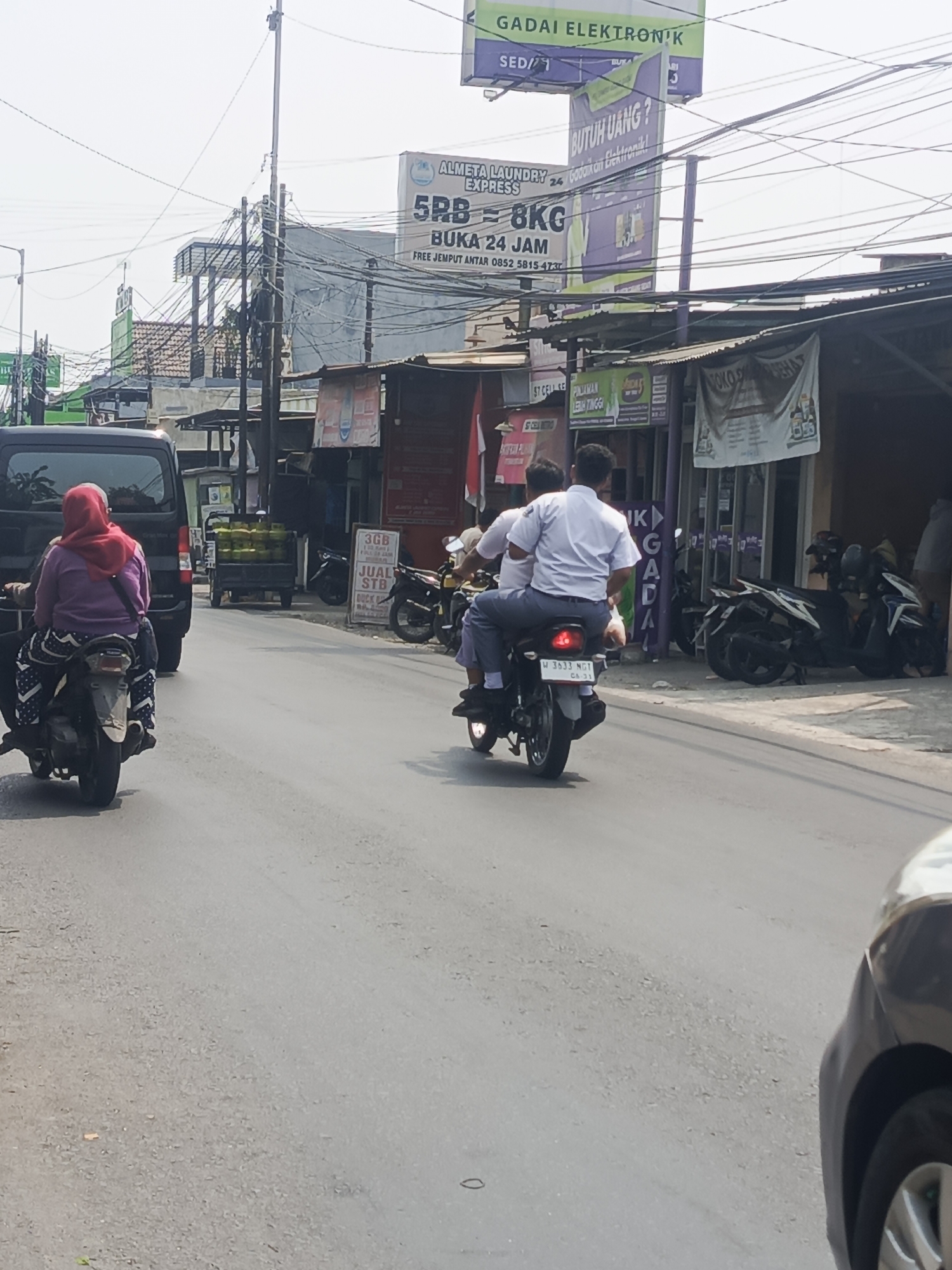 A street scene in Sedati Agung, Indonesia, features a busy road with several vehicles and buildings. In the foreground, a black car is partially visible on the right side of the frame. A motorcycle with two occupants, a woman in a pink hijab and a patterned skirt, and a passenger behind her, is moving away from the camera. The woman is wearing a pink hijab and a purple top. Further down the road, another motorcycle carries two young men, both wearing white shirts and blue pants, likely students. The background shows a variety of shops and buildings, including signs for "GADAI ELEKTRONIK" and "ALMETA LAUNDRY EXPRESS," indicating commercial activity. Overhead, a complex network of power lines crisscrosses the sky. The street is paved and appears to be a typical urban environment in Indonesia.