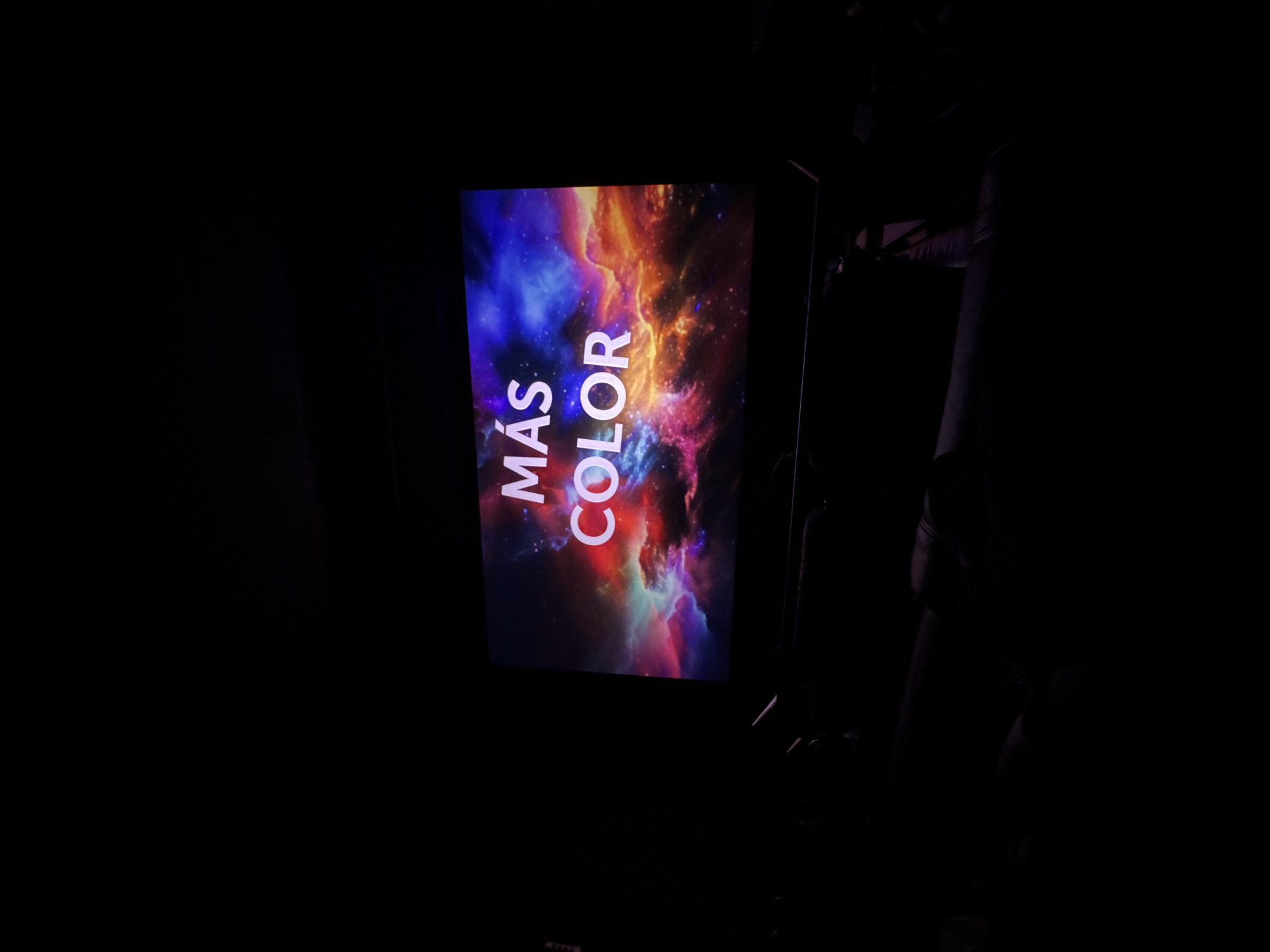 A screen in a darkened room displays a nebula-like image with the words "MÁS COLOR" vertically aligned. The image has a mix of blue, orange, and red hues. A stand or support structure is vaguely visible to the right of the screen. The overall environment appears to be a presentation or viewing setting. The city of Puebla, Mexico, is not discernible from the provided image.