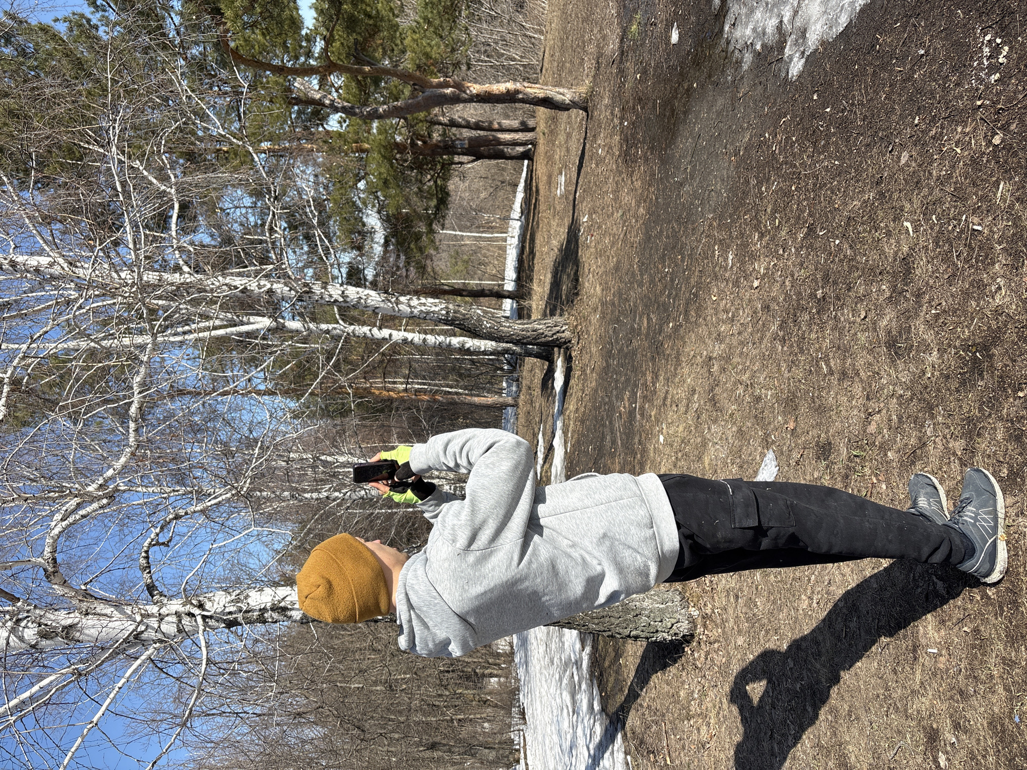 The image shows a person standing in a wooded area, holding a phone as if taking a picture. The person is wearing a mustard-colored beanie, a gray hoodie, black pants with cargo pockets, and gray running shoes. They are wearing gloves that appear to be neon green or yellow. The ground is a mix of dirt and what appears to be melting snow. Several bare trees are visible, along with evergreen trees in the background. The sky is blue. Based on visual cues and available information, the location is consistent with a park or natural area in Ulyanovsk, Russia.