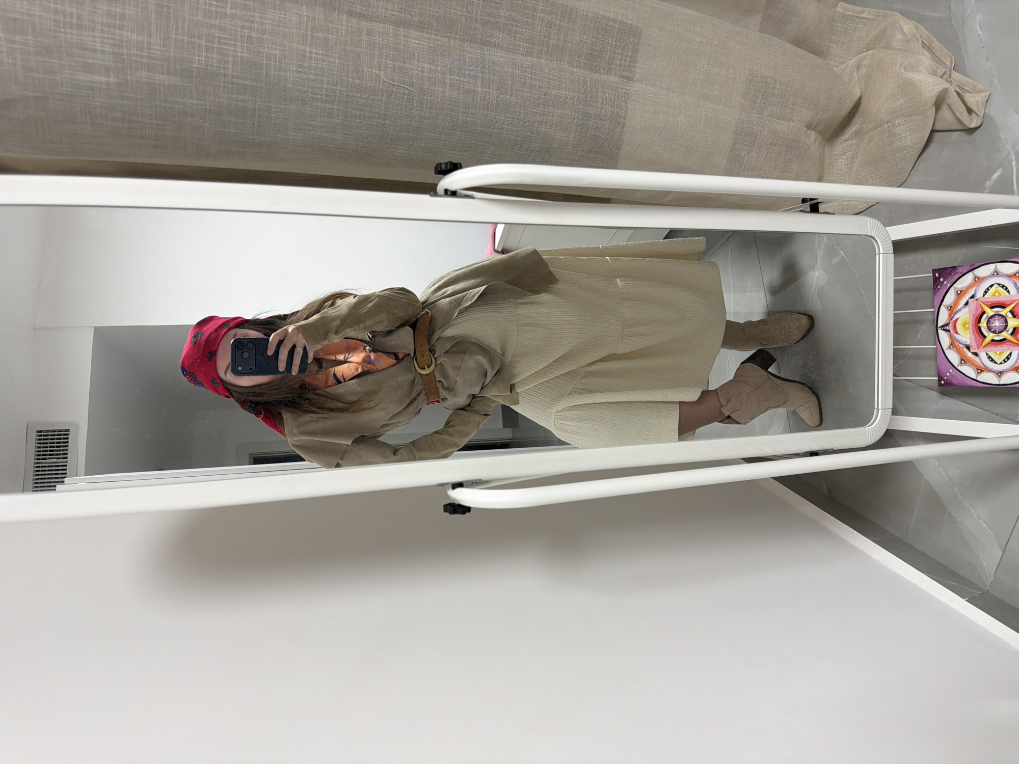 An image, originally rotated, displays a mirror selfie of a person's outfit in an indoor setting. The person's face is obscured by a smartphone, likely an iPhone, held up to take the photo, but a red headscarf with small patterns is visible. The outfit comprises a beige or khaki jacket or coat, cinched at the waist with a brown leather belt featuring a gold buckle, a long, light-colored, ribbed skirt or dress, and light brown ankle boots.
The mirror itself has a thin white frame and is part of a larger white stand. Visible outside the mirror's reflection, to the right, is a colorful circular artwork with a mandala-like design in shades of yellow, orange, red, and purple, set against a purple background. Above the mirror, a beige, sheer fabric or curtain is draped, and the floor is covered in gray tiles. A white wall with a vent is partially visible in the reflection to the upper left. A white surface or shelf extends into the foreground below the mirror. The scene suggests a casual fashion moment or outfit display. No legible text is visible in the image. The location context for the image is Bat Yam, Israel.
