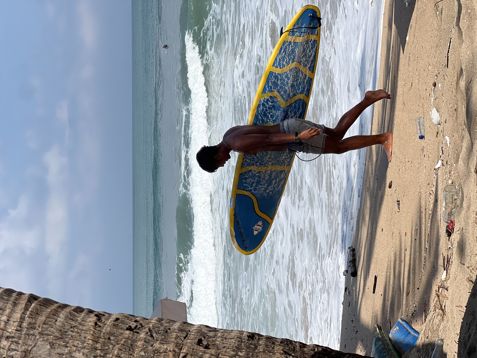 In Kuta, Indonesia, a young man with dark hair walks along a sandy beach carrying a blue and yellow surfboard. He is shirtless and wearing grey shorts. The ocean is to his right, with moderate waves breaking and a person on a surfboard in the distance. To the left of the frame, the rough bark of a palm tree trunk is visible. The beach is littered with debris, including plastic bottles, wood, and other small objects, with shadows cast by overhead foliage. The sky is overcast with a few patches of lighter blue.