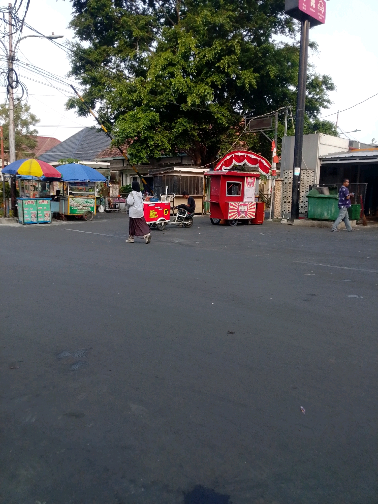 In East Jakarta, Indonesia, a street scene unfolds with several mobile food carts and a few individuals. The foreground is dominated by a gray asphalt road, with some faint markings. In the middle ground, a woman in a light-colored shirt and long brown skirt walks across the road. To her left, two food carts are set up under colorful umbrellas (yellow and red, and blue). A man appears to be working at one of these carts. Further to the right, a distinctive red food cart with a peaked red and white canopy and decorative trim is parked. This cart features signage with sunburst graphics and text that appears to say "KEBAB OLLIO." A person is seated on a motorcycle in front of this red cart. On the far right, a man in a blue patterned shirt and jeans walks past a large green dumpster. Behind the food carts, there are buildings with tiled roofs and a large, leafy tree. Overhead, a complex network of power lines and a street lamp are visible. The overall lighting suggests it is daytime, possibly late afternoon or early morning.