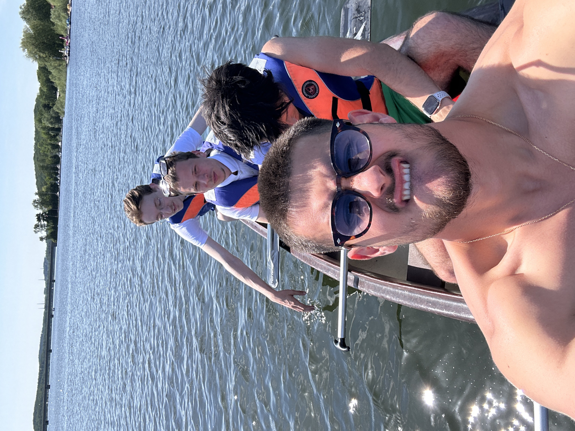 The image shows a group of four individuals on a body of water, likely the Möhnesee in Germany, based on the setting. The photograph appears to be a selfie taken by the person in the foreground, who is shirtless, wearing sunglasses and a gold chain. He has a beard and is smiling. Behind him, three other individuals are visible within the confines of a boat. They are wearing life vests. One person has dark hair. The boat is in the water, and one person is touching the water with their hand. The water is calm. Trees are visible in the background.