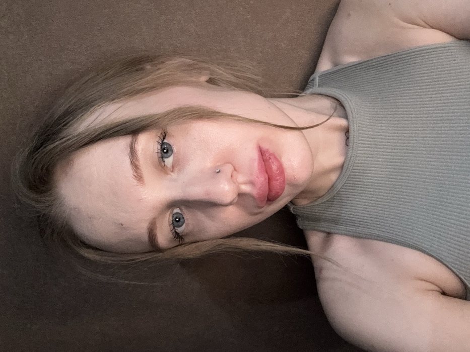 A close-up portrait shows a female subject with fair skin, light brown hair, and blue eyes, looking directly at the camera. She has a small metallic stud in her left nostril and her lips appear full with a reddish tint. She is wearing a light green ribbed tank top. Faint linear marks are visible on her left shoulder. A small dark tattoo is present on her upper sternum. The background is a plain, dark brown surface, out of focus. The scene takes place indoors in Rostov-on-Don, Russia.