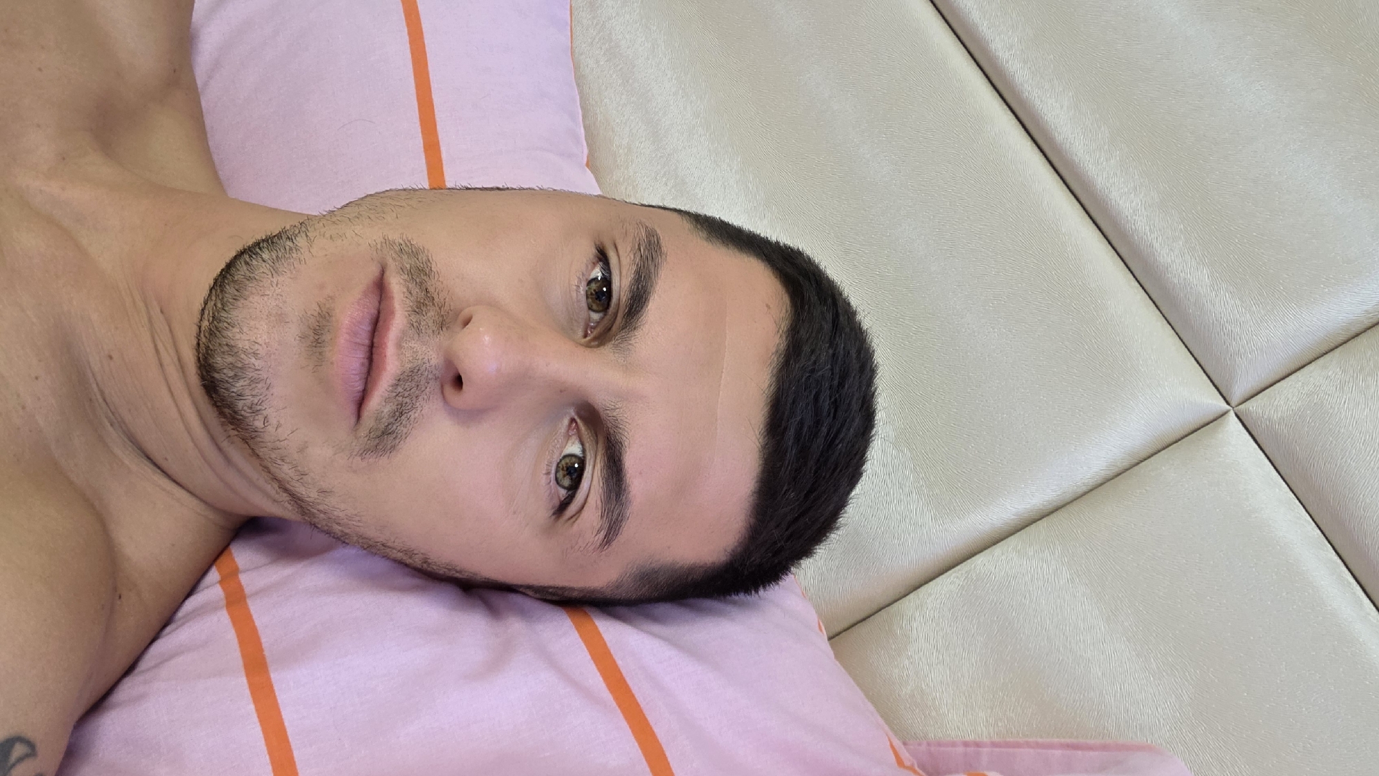 A close-up, high-angle, selfie-style photograph shows a man lying in bed, with his head resting on a pink pillow with orange stripes. The background consists of a padded headboard with a diamond pattern. The man has short, dark hair, brown eyes, and a light beard and mustache. He is looking directly at the camera. The lighting suggests an indoor setting, possibly during the day. There are no visible indications of the location being in Blagoevgrad, Bulgaria; the scene is generic for a bedroom.