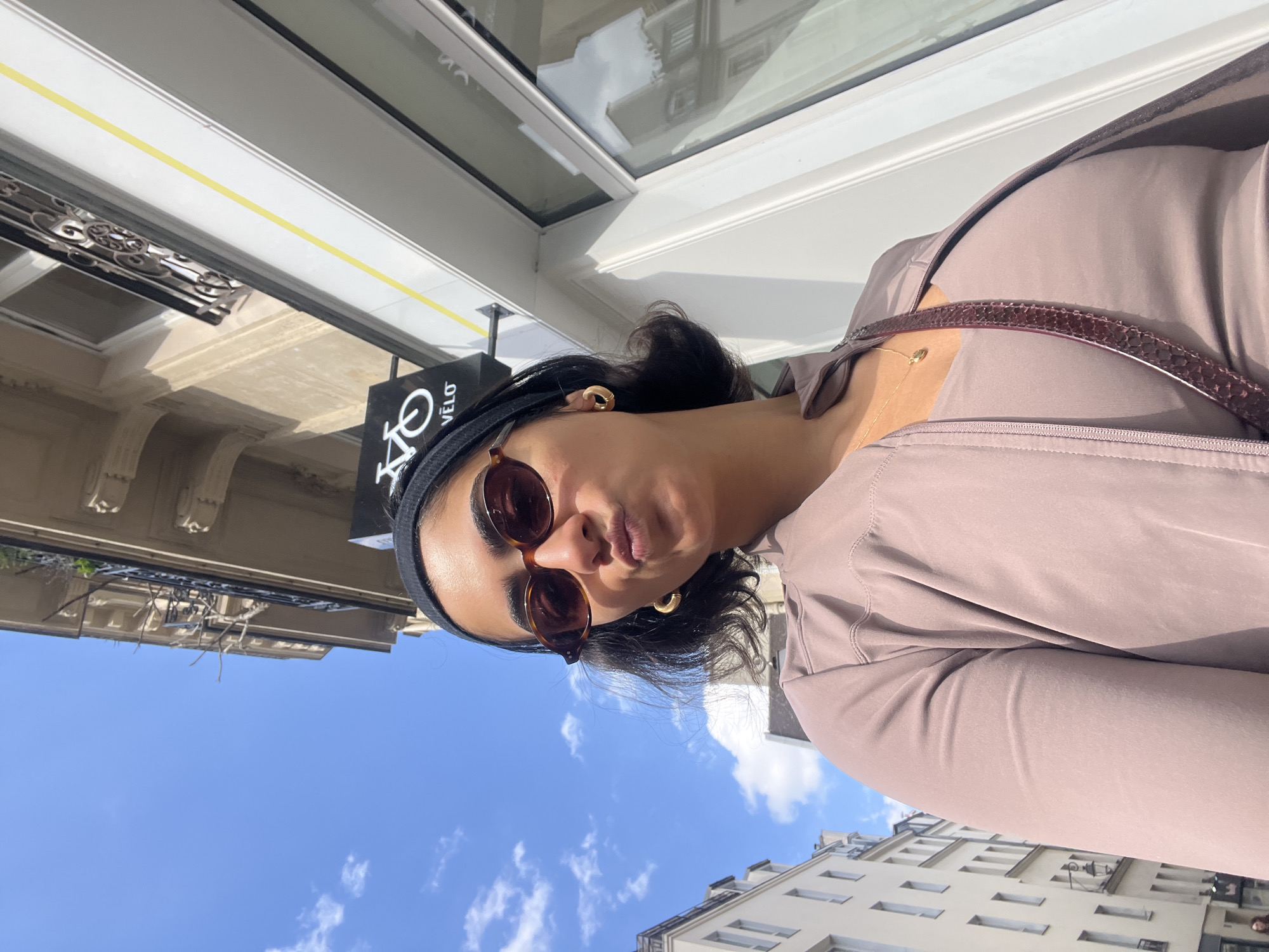A medium shot captures a woman in Paris on a sunny day. She wears a headband, sunglasses, gold earrings, and a light purple athletic zip-up jacket with a crossbody bag. The background features a building with white window frames, reflecting the sky, and a black sign with white lettering that reads "VÉLO." Behind her, the blue sky is visible, dotted with clouds, and below the sky, a building with repeating windows is seen. The architectural style and window patterns are characteristic of Parisian buildings.