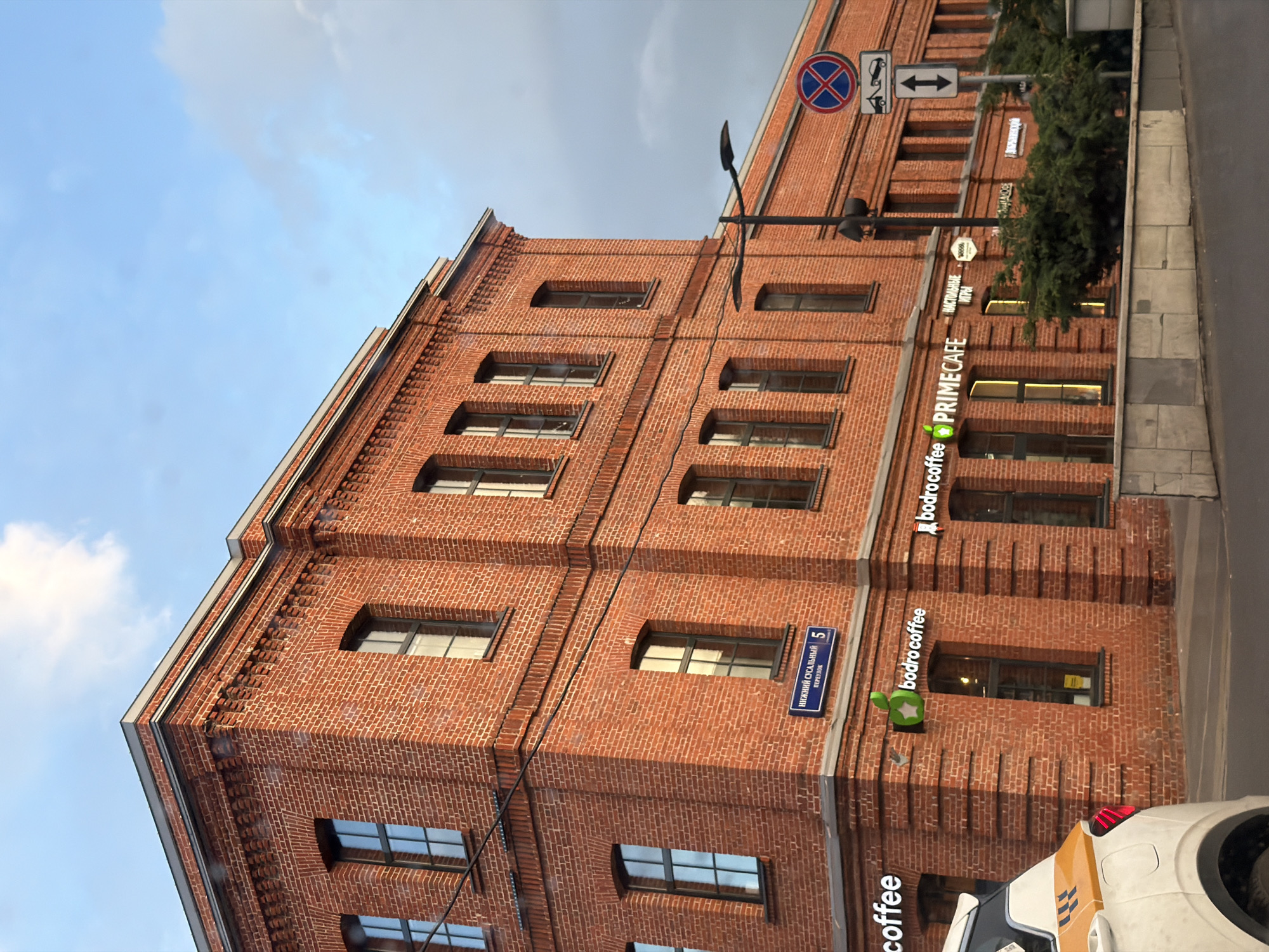 The image shows a red brick building. On the building there are signs for "bodro coffee" and "PRIMECAFE". There is a street sign that reads "Нижний Сусальный переулок 5", which translates to "Nizhny Susalny Pereulok 5". Based on the street name, the image is likely taken in Moscow, Russia. There is a car parked on the right side of the image. The sky is visible in the upper left corner.