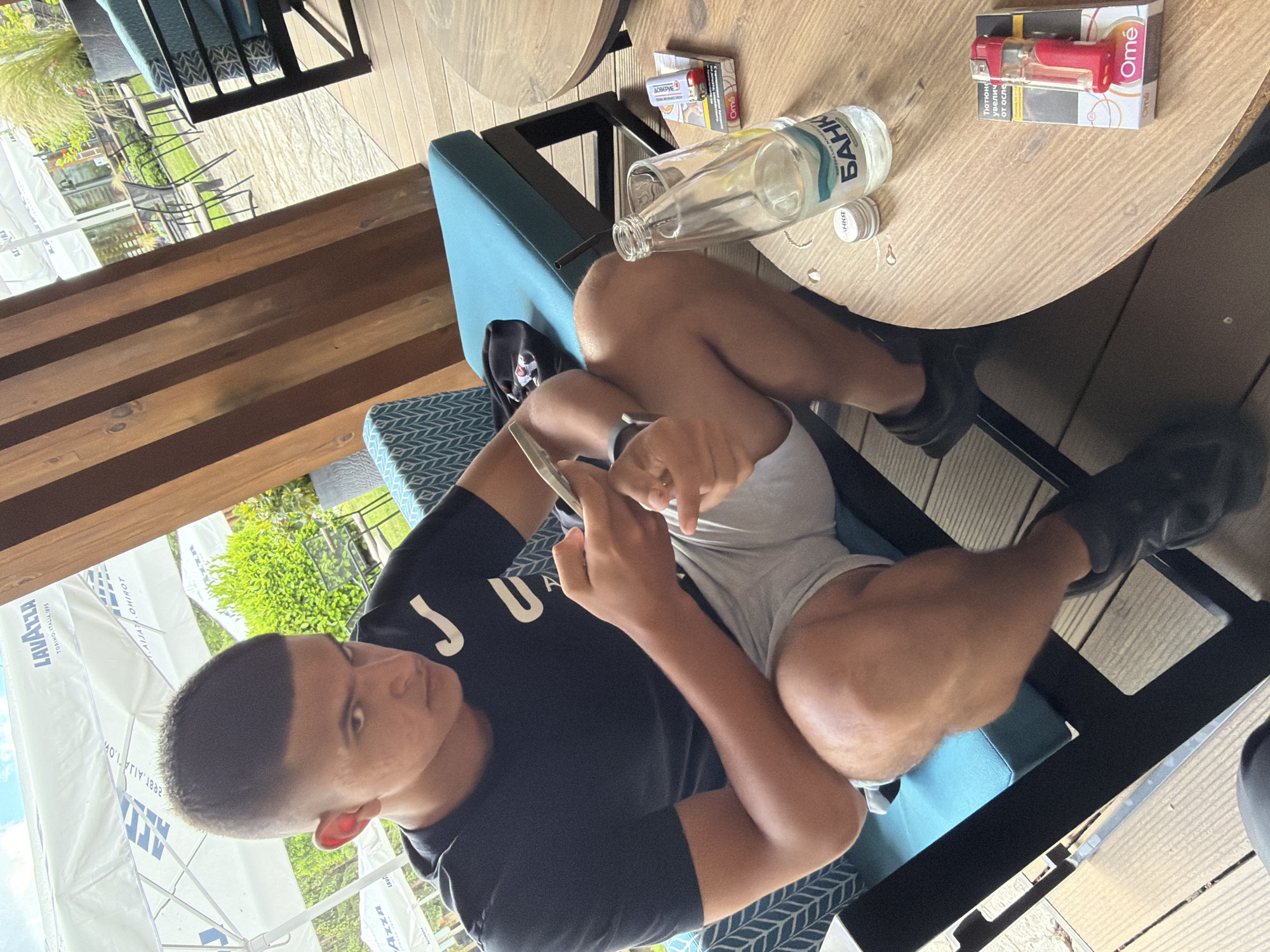 A young man is depicted in a casual outdoor setting, likely a cafe or a relaxed patio, in Razlog, Bulgaria. He is seated on a blue-patterned chair, leaning back while engaged with a smartphone held in his hands. He wears a black t-shirt, which partially displays "JUA" as part of a larger, obscured word, and light-colored shorts with black shoes.
Adjacent to him, on a round wooden table, are several items. These include an empty glass bottle of "БАНКЯ" (Bankya) brand water, along with its white cap. Next to the bottle, a packet of "Omé" cigarettes is visible, topped with a red and clear plastic object that appears to be a lighter or a vape device. Partial Bulgarian text on the cigarette packet reads "Тютюно увелич от осле", likely a health warning.
The scene is illuminated by bright daylight, suggesting a sunny day. In the background, a large white umbrella with the "LAVAZZA TORINO, ITALIA, 1895" logo provides shade. Other outdoor furniture, such as additional chairs and tables, and some green foliage are discernible, indicating an open-air environment. The flooring is made of wooden decking. The overall impression is one of leisure and relaxation. The image itself is captured with a tilted perspective.