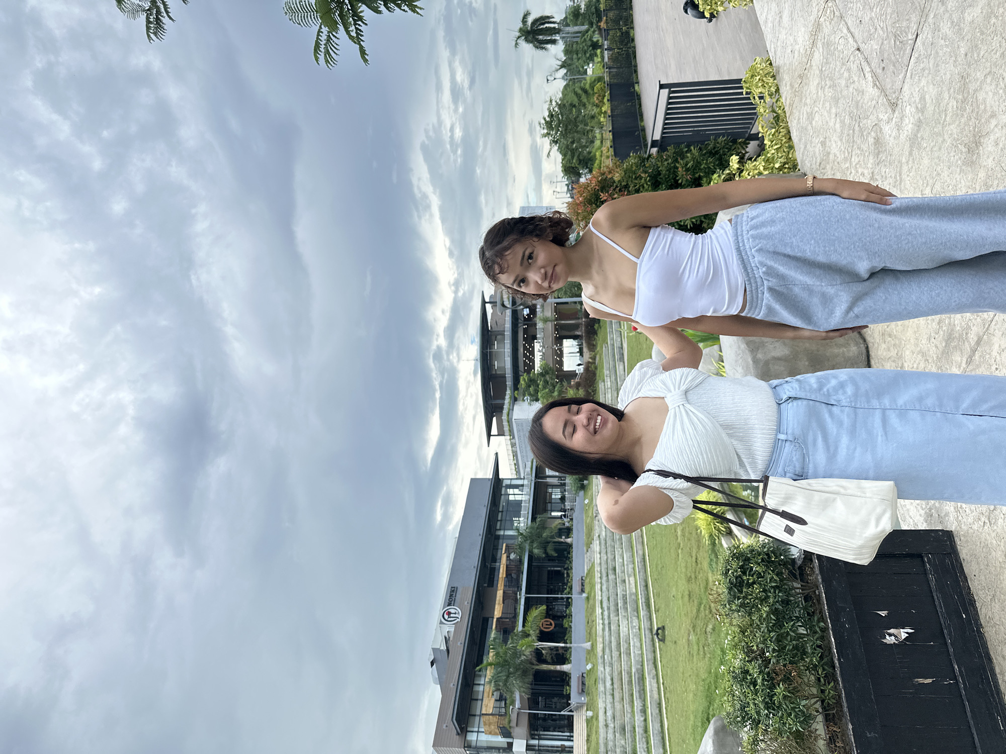 Two young women are standing outdoors in front of a modern building complex, with a cloudy sky overhead. The woman on the left is wearing a white off-the-shoulder top with a knotted detail at the bust and a light blue denim skirt. She is carrying a white tote bag with dark brown straps over her left shoulder and has her right hand raised, touching her neck. She is smiling with her eyes closed. The woman on the right is wearing a white tank top and light grey sweatpants. She is standing with her arms at her sides and is looking towards the camera. Behind the women, there is a tiered grassy area leading up to a multi-story building with large windows. The building has a sign that reads "MONKI." To the right of the building, there are more structures, trees, and a railing. The foreground consists of a paved walkway and some planters with greenery. The overall setting appears to be a commercial or recreational area in a city. This scene is consistent with locations found in Davao City, Philippines, likely a modern development with commercial establishments and outdoor spaces designed for public use.