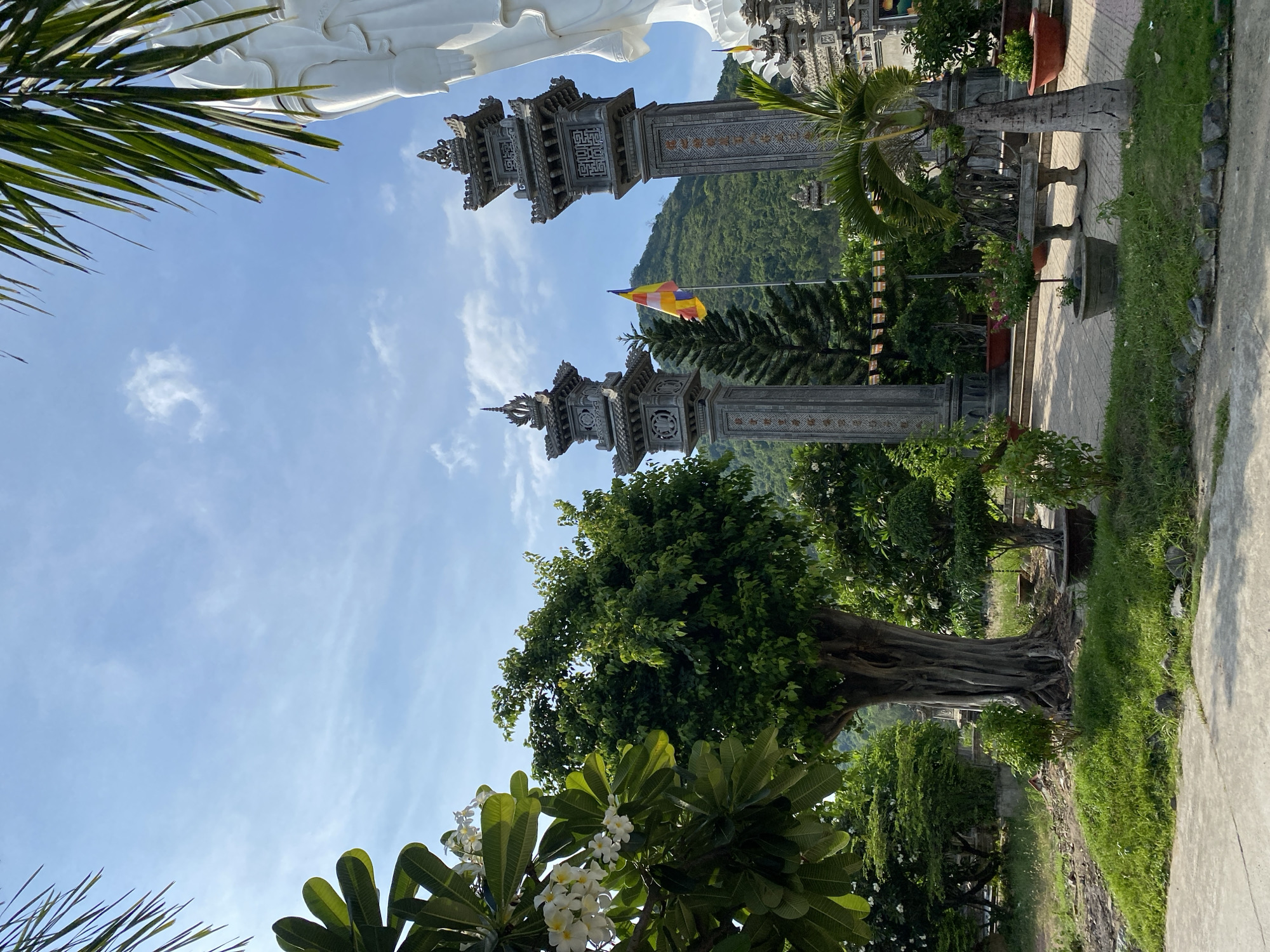 The image depicts an outdoor scene at a temple complex in Tay Nha Trang Ward, Vietnam. In the foreground, lush green grass covers the ground, bordering a paved pathway. A large, ancient tree with gnarled roots stands prominently to the left, its branches laden with green leaves and white flowers. To the right, a young palm tree with a slender trunk is visible.
Dominating the midground are two ornate, grey stone pillars, each topped with intricate pagoda-like structures. Between these pillars, a multi-colored flag with horizontal stripes of yellow, red, and blue flutters in the breeze. Behind the pillars, a majestic white statue of a deity, partially visible from the shoulder up, rises against the sky. The background features a verdant, tree-covered mountain range under a clear blue sky with scattered white clouds. The overall impression is one of tranquility and spiritual serenity, enhanced by the natural beauty and architectural elements of the temple.
