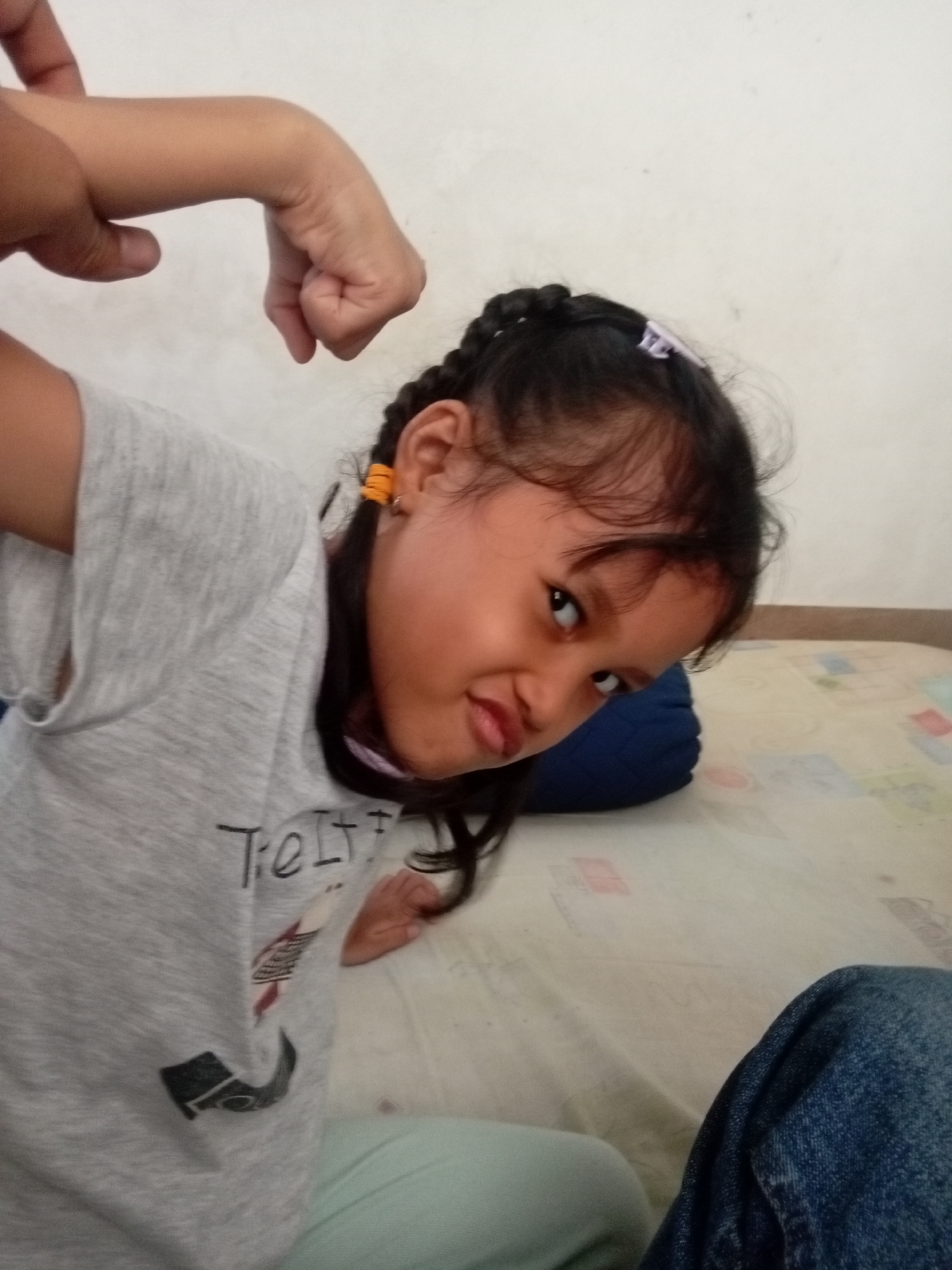 In Batam, Indonesia, a young girl with dark hair styled in braids and adorned with small yellow earrings, sits on a white mattress. She is wearing a light grey t-shirt with a graphic print and light green pants. Her left arm is flexed, with her hand forming a fist, as she leans her body forward and turns her head to look at the camera. Her facial expression is playful, with her mouth slightly open and her eyes looking directly at the viewer. Behind her, a dark blue pillow is visible. The background is a plain, light-colored wall. A portion of dark blue denim pants is visible in the lower right corner of the frame. The lighting suggests an indoor setting, possibly a bedroom.