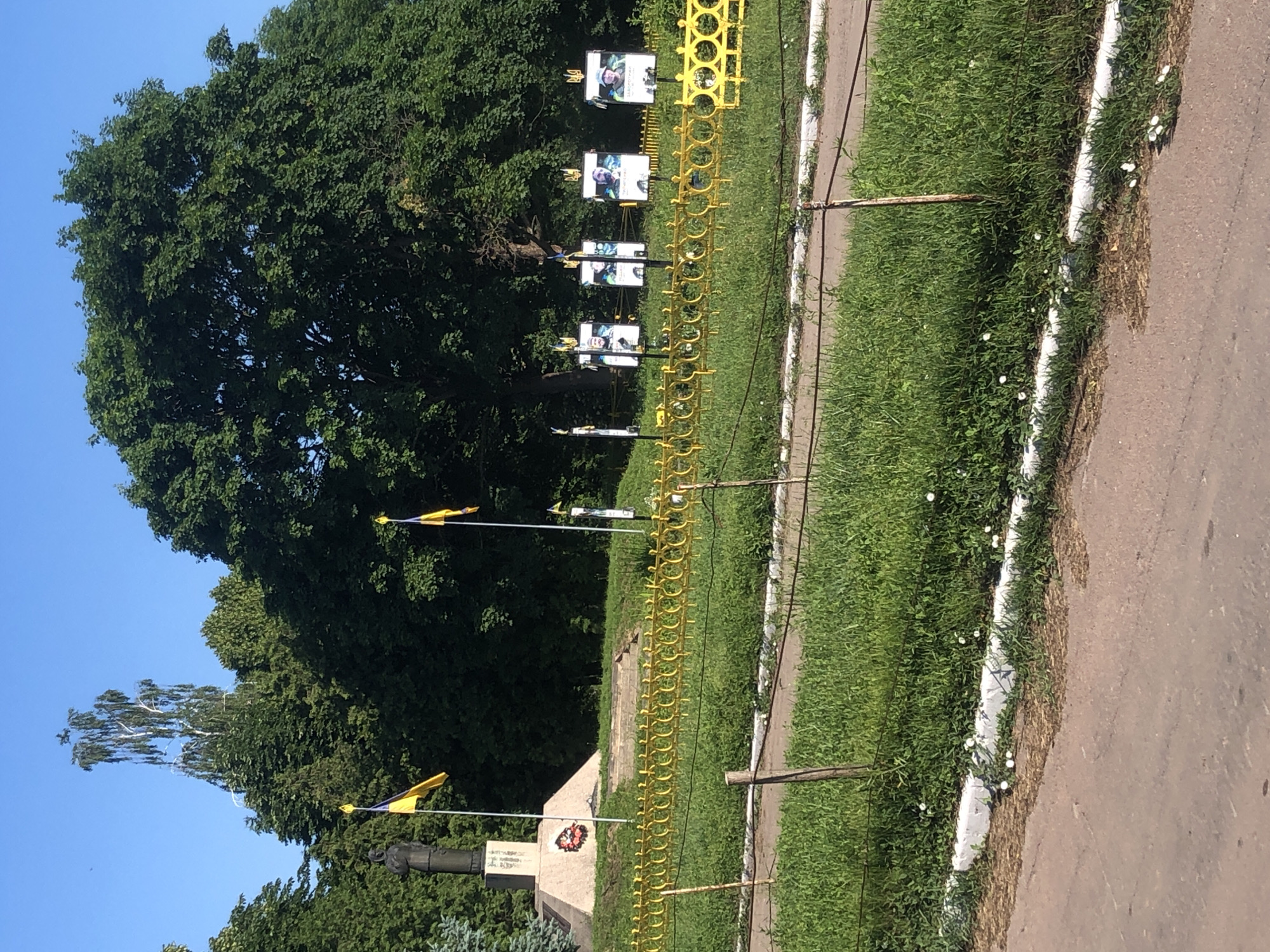 The image shows a monument with two Ukrainian flags in front of a large tree. There are also four framed photographs of people displayed on poles. In the foreground, there is a yellow decorative fence and green grass. The monument is located in Khmilnytsia, Ukraine. The monument appears to be a statue on a pedestal. The photographs show individuals, likely soldiers or heroes, with Ukrainian flags and symbols. The environment is outdoors with clear blue sky and lush green trees.