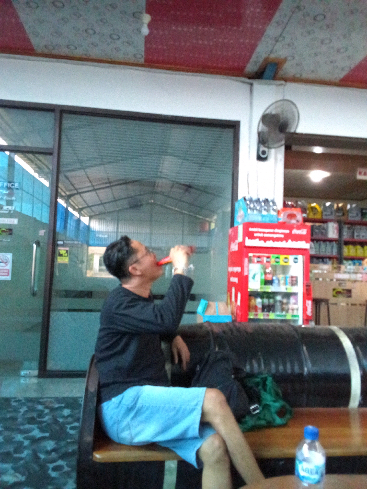 The image depicts a man in the city of Badas, Indonesia, seated on a bench and drinking from a red object. He is wearing a black long-sleeved shirt and light blue shorts. To his left, a glass door with "OFFICE" signage is visible. Behind him, a red beverage cooler is stocked with drinks. The environment appears to be an indoor waiting area or a shop.