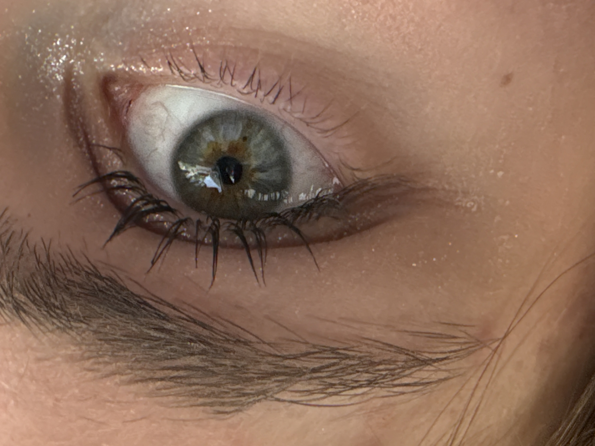 A close-up image shows a human eye with long, dark eyelashes. The iris is multi-toned with grey, green, and brown segments. There are prominent blood vessels in the sclera. There is a reflection on the pupil. An eyebrow is visible below the eye. The surrounding skin has a light, fair tone, and there are traces of shimmer makeup on the eyelid. A small mole is visible above the eye. The image does not contain sufficient information to determine if the photograph was taken in Perm, Russia.