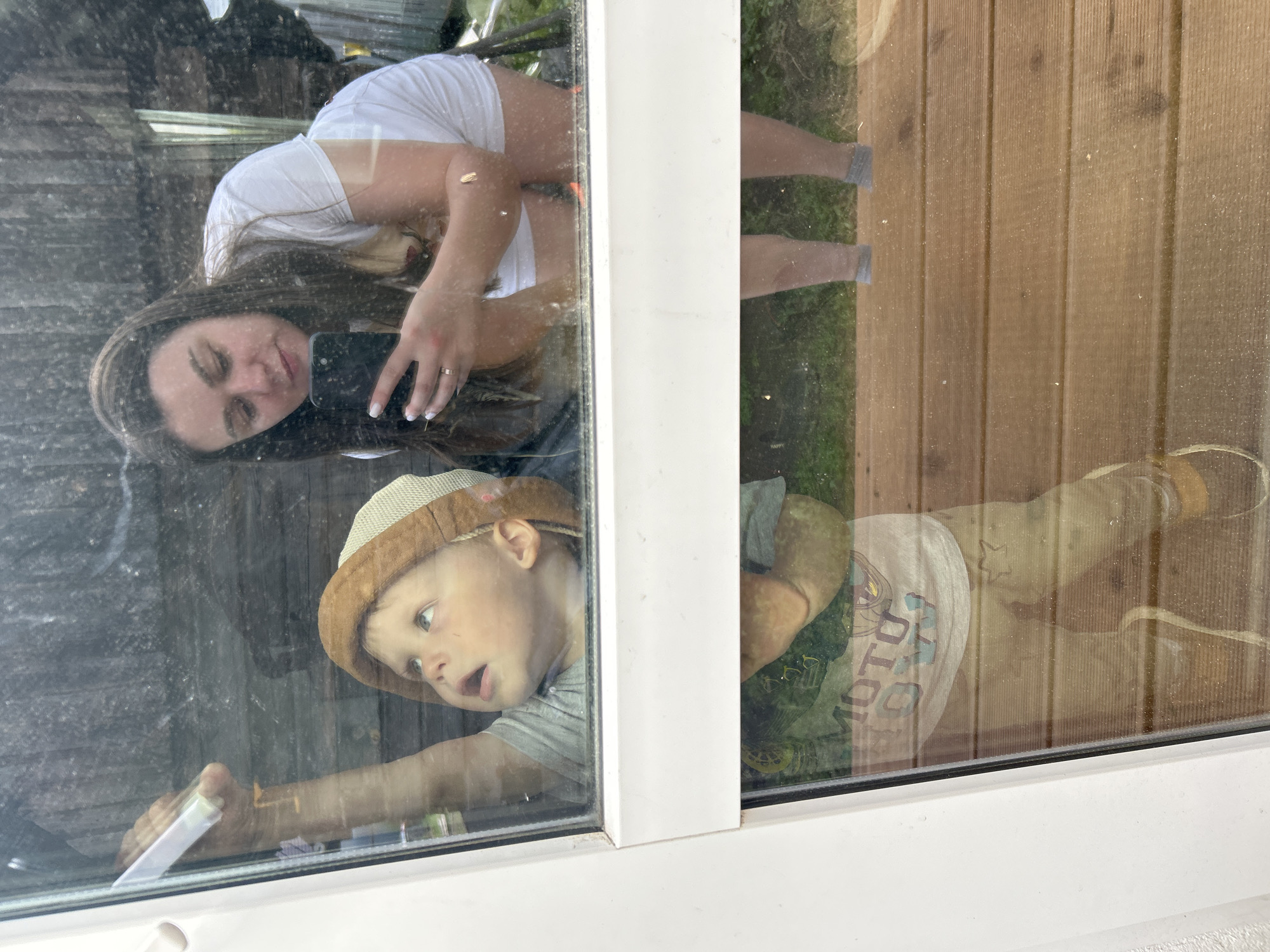 A child wearing a bucket hat and grey t-shirt is looking up and to the left, his right arm extended upward, holding a white object. A woman with long brown hair, wearing a white t-shirt and white shorts, is reflected in the glass, holding a phone and taking a selfie. The reflection shows her looking down at the phone. The background reflected in the glass appears to be an outdoor setting with wooden planks, possibly a deck, and some greenery. The overall scene is captured through a dirty window, with the dirt obscuring some details of the background and reflections. There is no information in the image that can be used to determine the location in the city of Chaikovsky, Russia.