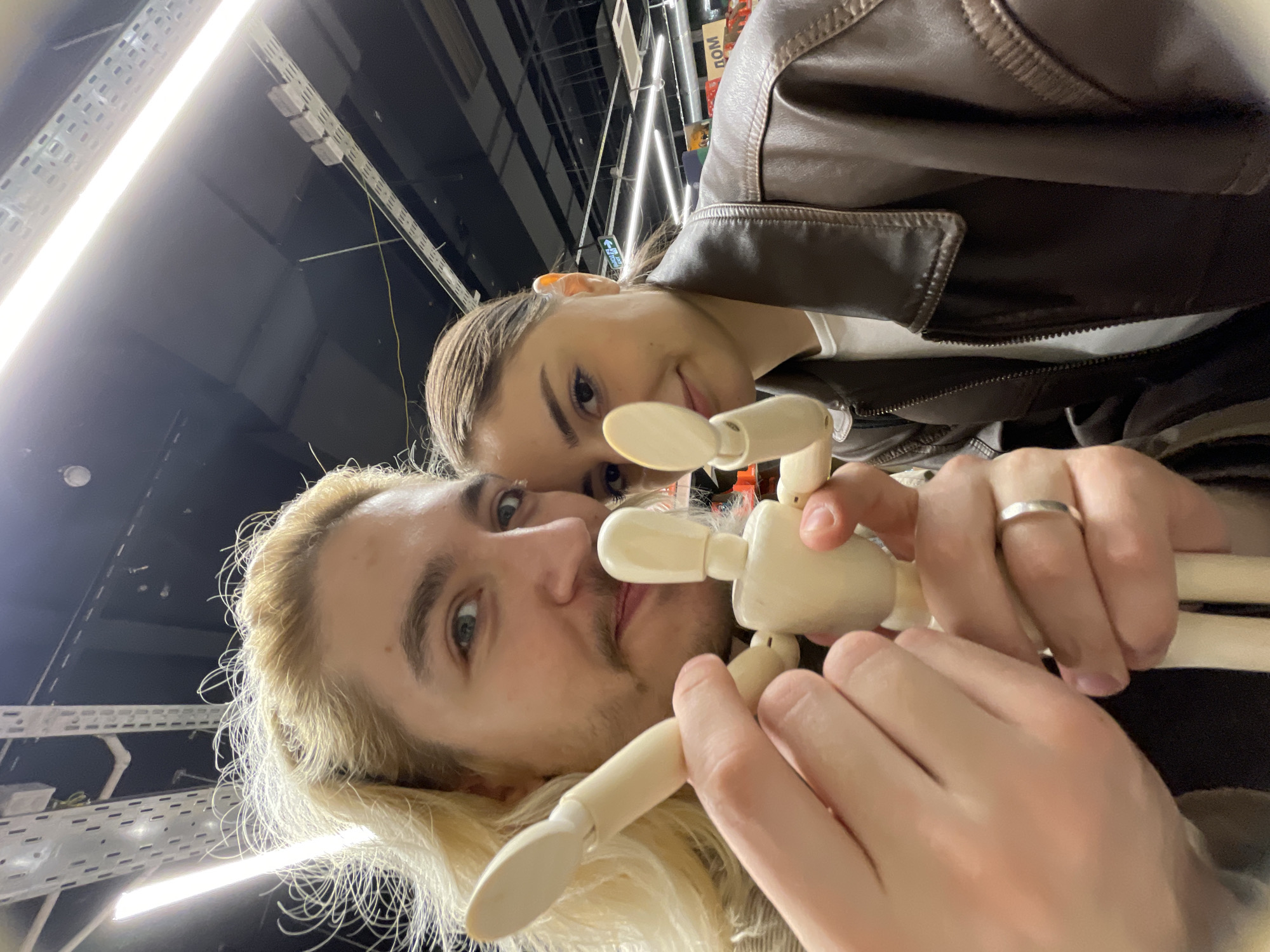 The image shows a selfie taken from a low angle, looking up. Two individuals are present. The person in the foreground has blond hair and is holding a wooden articulated mannequin. Their hands are visible, with a ring on one finger. Behind them, a woman with her hair pulled back is smiling at the camera. The background shows a store interior with overhead lighting and rows of shelves. Based on the text visible in the background, it appears to be a retail establishment in Moscow, Russia. The foreground person is wearing a brown leather jacket.