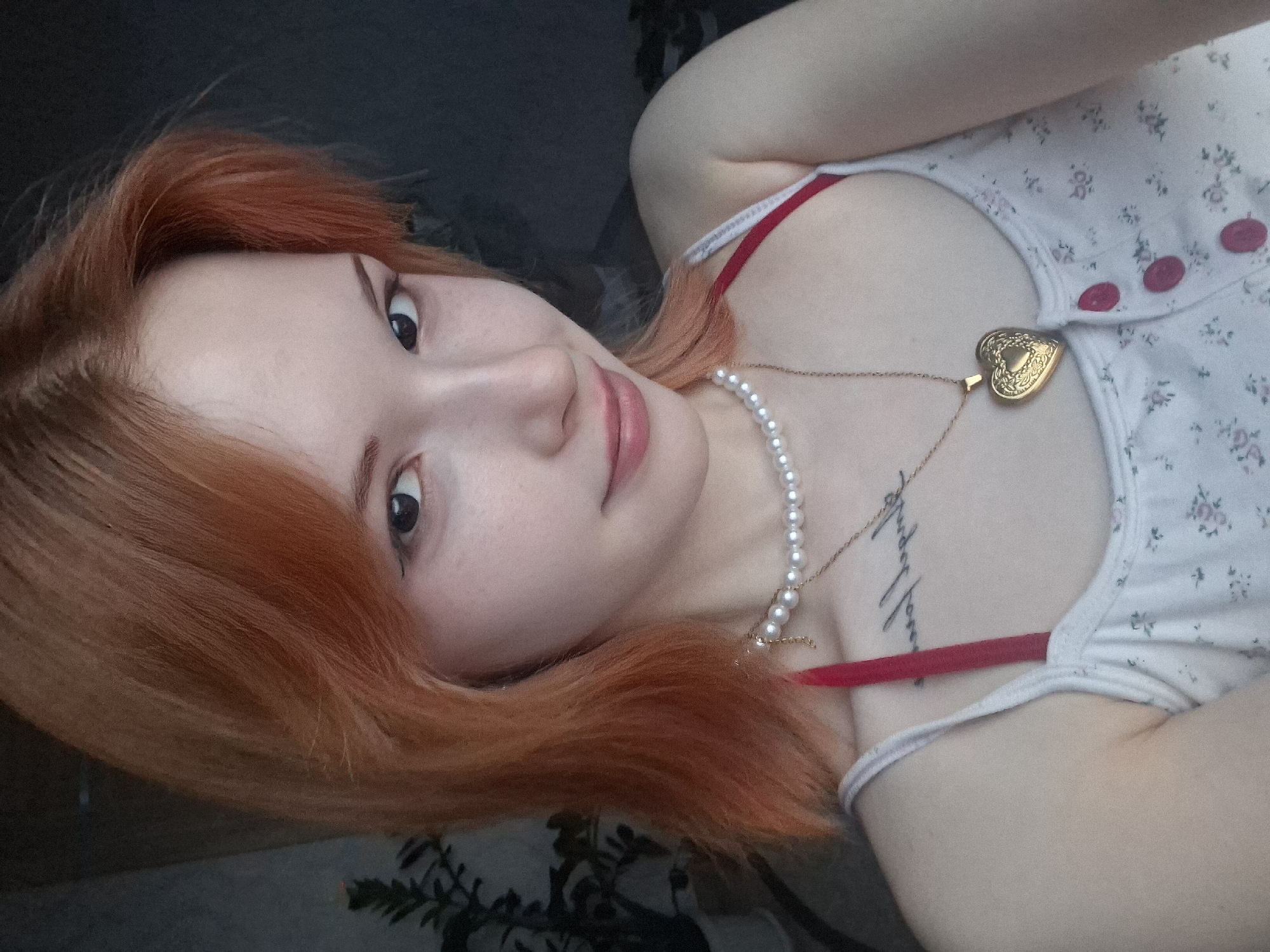 A close-up, rotated portrait features a young individual, likely female, with fair skin and vibrant reddish-orange hair. The person is positioned horizontally in the frame, looking directly at the camera with dark eyes and a subtle, gentle smile. Their facial features suggest youth, with natural-looking makeup.
They are wearing a light-colored top, possibly white or cream, adorned with small, delicate floral patterns in muted colors and three distinct pink buttons on the front closure. A red bra strap is visible beneath the top's strap on the person's left shoulder (right side of the image). Adorning their neck are two necklaces: one a strand of lustrous white pearls, and the other a thin gold chain with a detailed, heart-shaped gold locket as a pendant. A tattoo, featuring illegible text in a script font, is visible on their upper chest, beneath the necklaces.
The background is dark and out of focus, suggesting an indoor setting. A blurred green plant or foliage is visible in the lower right corner of the image. The overall impression is one of calm and direct engagement with the viewer. This image was taken in Chaikovsky, Russia.