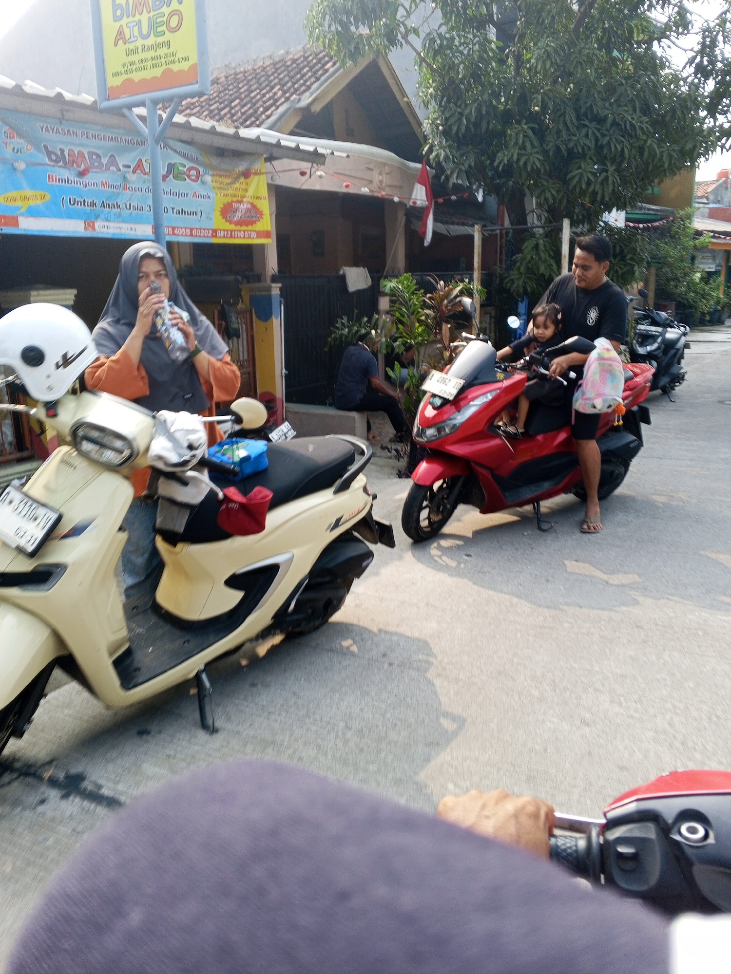 The image depicts a street scene in Ciruas, Indonesia, possibly in front of a building with commercial signage. In the foreground, a light-colored scooter is parked, and a person wearing a grey hijab and an orange top is holding and drinking from a bottle. To the right, a red scooter is occupied by a man in a black t-shirt holding a young child on his lap. The child is wearing a light-colored top and has their hair in pigtails. The man is looking towards the child. The background shows buildings, trees, and other parked motorcycles. Shadows of people and objects are cast on the asphalt ground. The overall atmosphere appears to be daytime, with sunlight illuminating the scene.