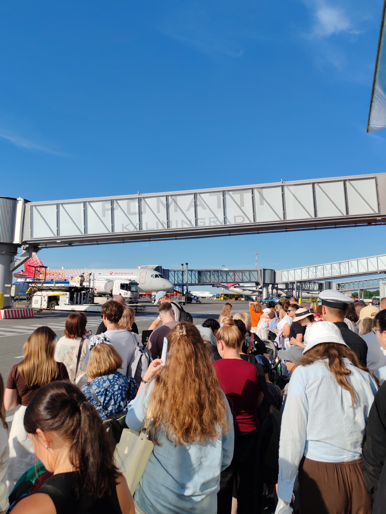 The image depicts an outdoor scene at an airport, likely Khrabrovo, Russia, given the context. A group of people are standing in what appears to be a queue, facing away from the camera. They are gathered on the tarmac, with an airplane and a jet bridge visible in the background. The weather appears to be clear and sunny, indicated by the bright blue sky and the shadows cast on the ground.
The people are dressed in casual summer clothing, suggesting a warm time of day. Some individuals are carrying bags, and one person in the foreground is wearing a white hat. The overall mood seems neutral, with no strong emotions readily apparent.
In the background, a "ROSSIYA" branded airplane is parked. A jet bridge connects to the terminal, and other airport vehicles and infrastructure are visible, including a tow truck with the number "28" displayed on its side, and a refueling truck. The jet bridge has text printed on it, which appears to be "PALMANTI KALININGRAD". The scene is set during the day, with ample sunlight.