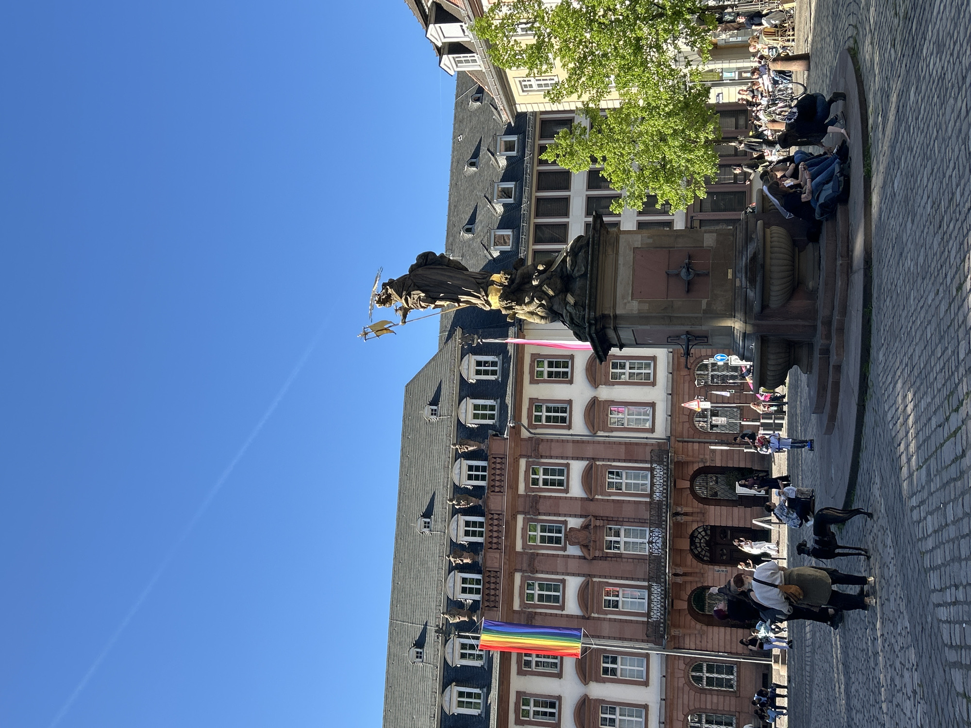 Captured on a sunny day, the photograph displays a city square. In the foreground, a statue crowns a fountain with figures clustered around its base, a few people lounging on the fountain's edge. A rainbow flag flutters from a building's facade, its architecture a mix of dark slate roofing and light-colored walls with dark accents. People populate the square, some sitting, some walking. The cobblestone ground adds texture to the scene. The sky is a clear, vibrant blue. The location is identified as being in the city of Unknown, Unknown.