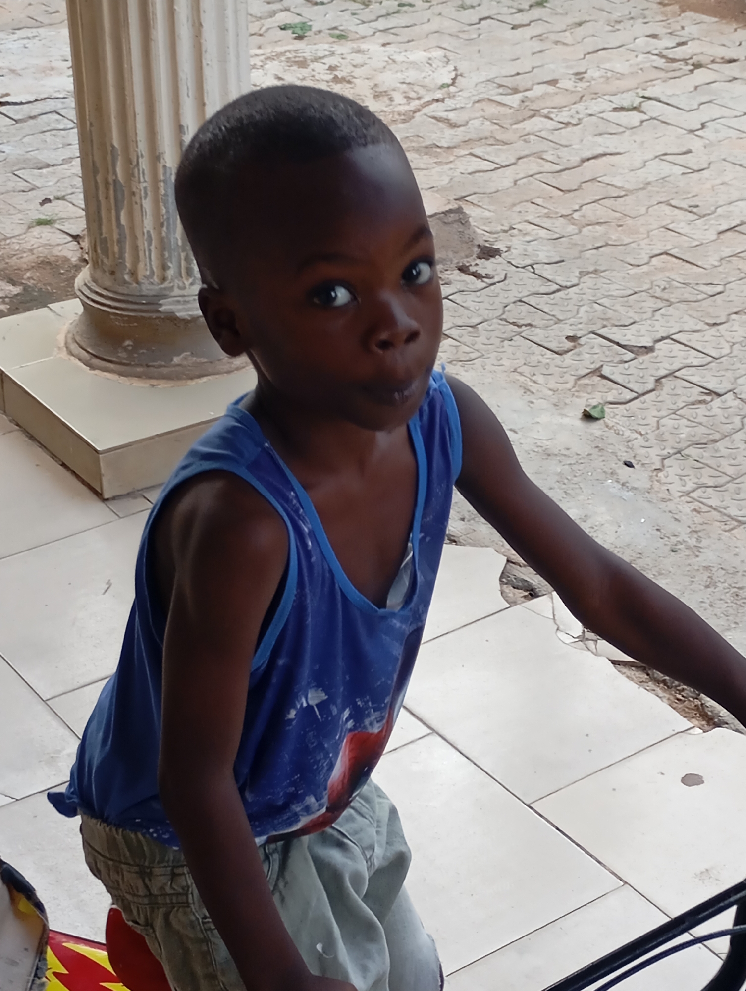 A medium shot shows a young, Black boy in a blue tank top standing outdoors in an unknown area of an unidentified city. The boy looks upward toward the camera with wide eyes and a slightly pursed mouth. He wears light-colored shorts. The location appears to be a covered patio or porch area with tiled flooring. A white column is visible to the left of the boy. A patterned, interlocking brick walkway is partially visible in the background. Part of a bicycle is visible at the bottom of the frame.