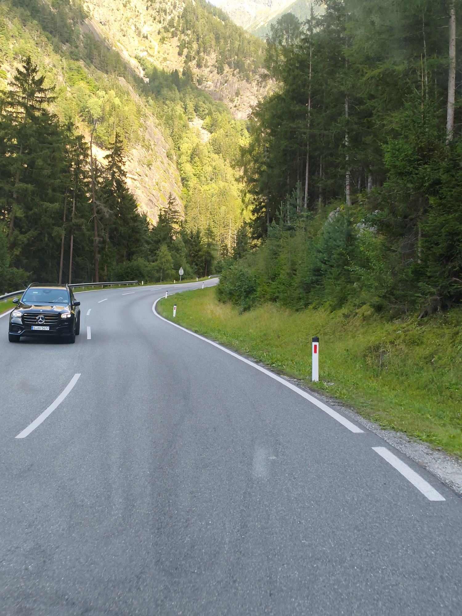 A black Mercedes-Benz SUV is driving on a winding asphalt road through a mountainous forest landscape. The car is a Mercedes-Benz GLB, based on its front grille and headlight design. The license plate is visible and reads "S-AV 5677". The road is marked with white dashed lines and has a white line on the right shoulder. There are several white posts with red tops along the right side of the road, indicating the edge of the pavement. The surrounding environment is characterized by dense green coniferous forests on a steep, rocky mountainside. Sunlight is illuminating the upper parts of the mountain, creating a contrast with the shaded forest. The sky is not visible in the frame. The image appears to be taken from a moving vehicle, likely following the SUV. The location is identified as being on the Ötztaler Straße (B186) in Oetz, Austria.