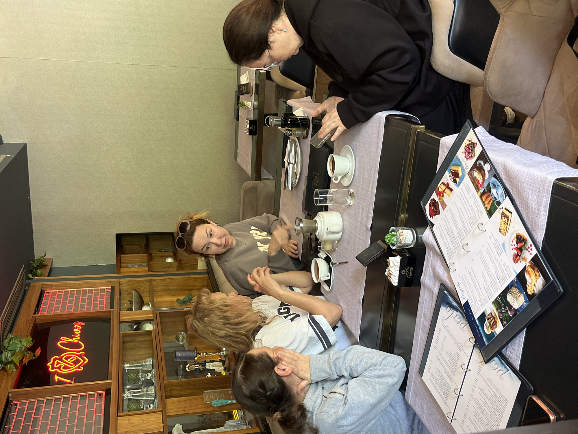 The image shows four women at a table in a restaurant. Three of the women are seated at the table, examining menus and engaging in conversation, while the fourth woman stands and appears to be taking a photograph with her mobile phone. The table is set with coffee cups, glasses, a coffee press, and dining utensils. There are two menus on the table.
The restaurant's interior features a wooden shelf unit with various decorative items, including a red neon sign. The setting appears to be "Perla," a restaurant or café located in Budva, Montenegro.
