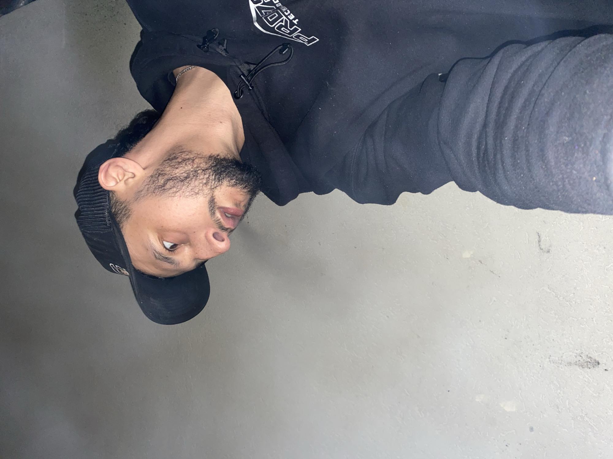 A man wearing a black baseball cap and a black hoodie with white lettering is angled in the upper left quadrant of the image, while a gray concrete surface fills the rest of the frame. The man has dark hair and a beard. He is wearing a silver chain around his neck. The lettering on the hoodie says "PROVZ TECH." The location is indiscernible in the image.