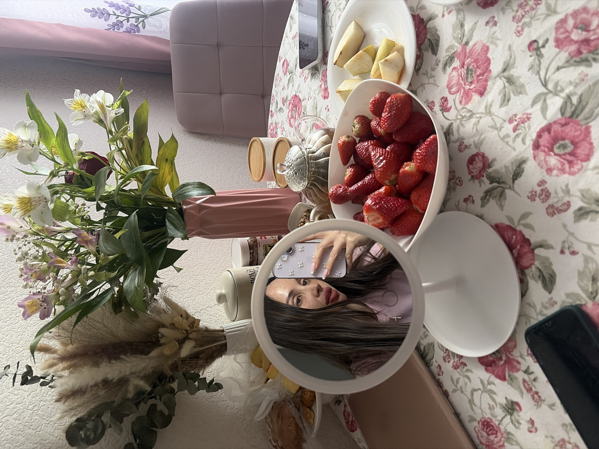 The image depicts a scene on a table covered with a floral tablecloth, likely indoors, although the location in Saint Petersburg, Russia, is not directly discernible from the visual content. On the table, there's a bowl of strawberries, a plate with cut pears, a teapot, and a vase with flowers. A mirror reflecting a person holding a phone is also visible, partially obscuring the view. The phone in the mirror appears to be used to take the picture, as the person in the reflection is holding the phone up. Other objects include jars and a bed headboard in the background. The overall scene suggests a casual, personal setting, possibly a breakfast or brunch arrangement.